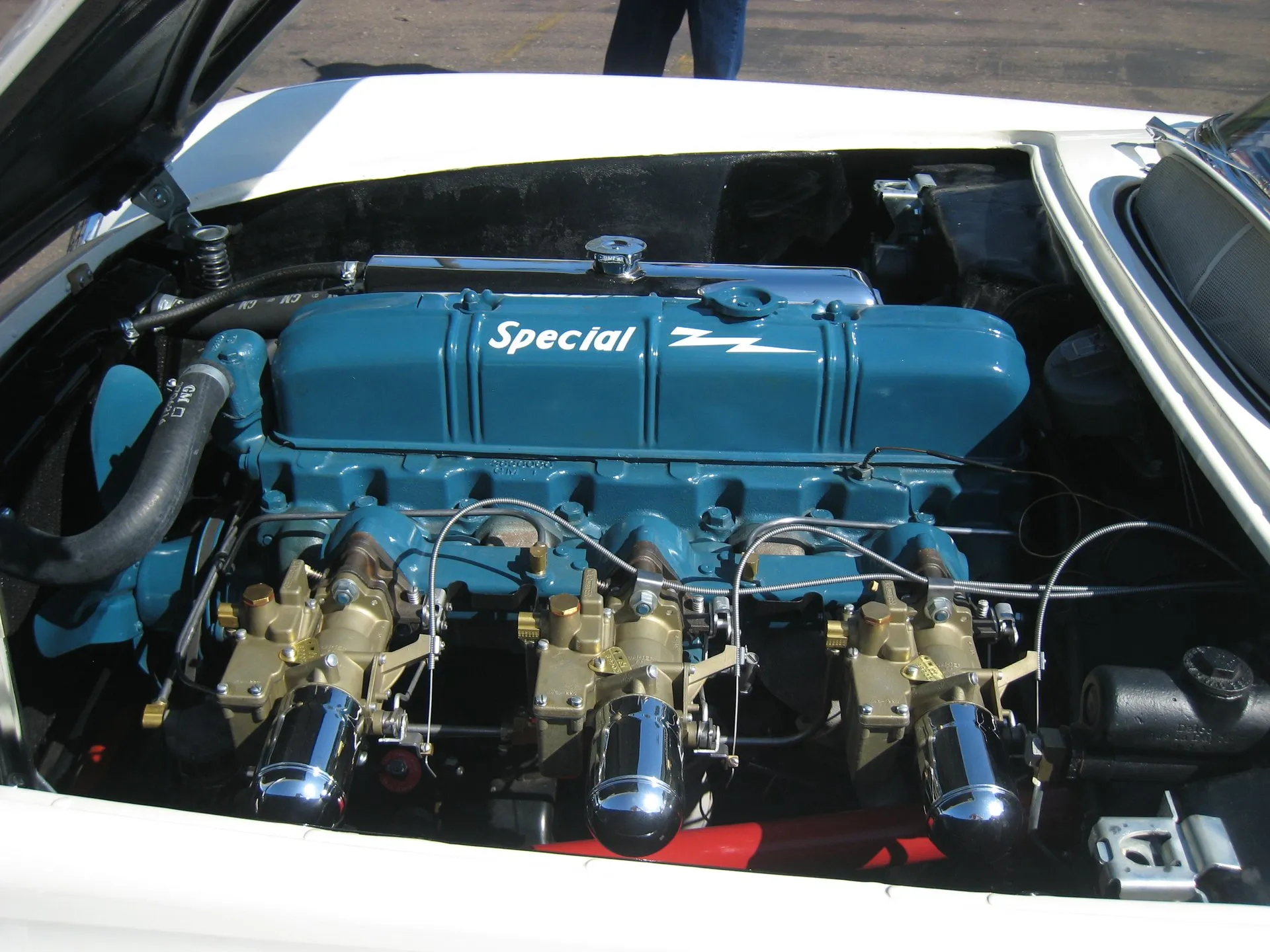 The open engine bay of a white 1953 Corvette: a blue inline six with Special script on its valve cover and three brass-colored carburetors topped by chrome bullet-shaped air cleaners.