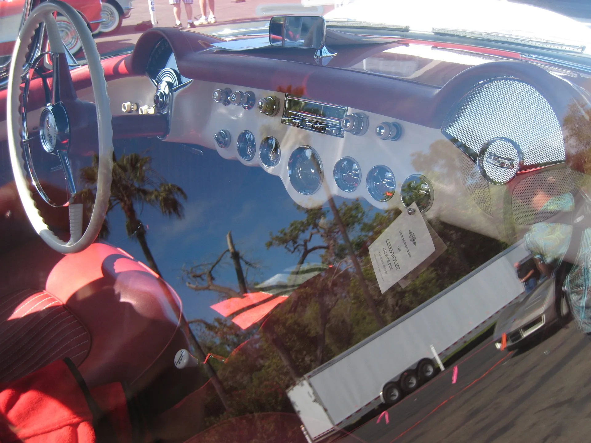 The cockpit of a 1953 Corvette seen from the driver's side: a white steering wheel, a red seat, a row of round chrome-ringed gauges across a white dash with a radio above them and a speaker grille on the passenger side, with palm trees and a trailer reflected across the glass.