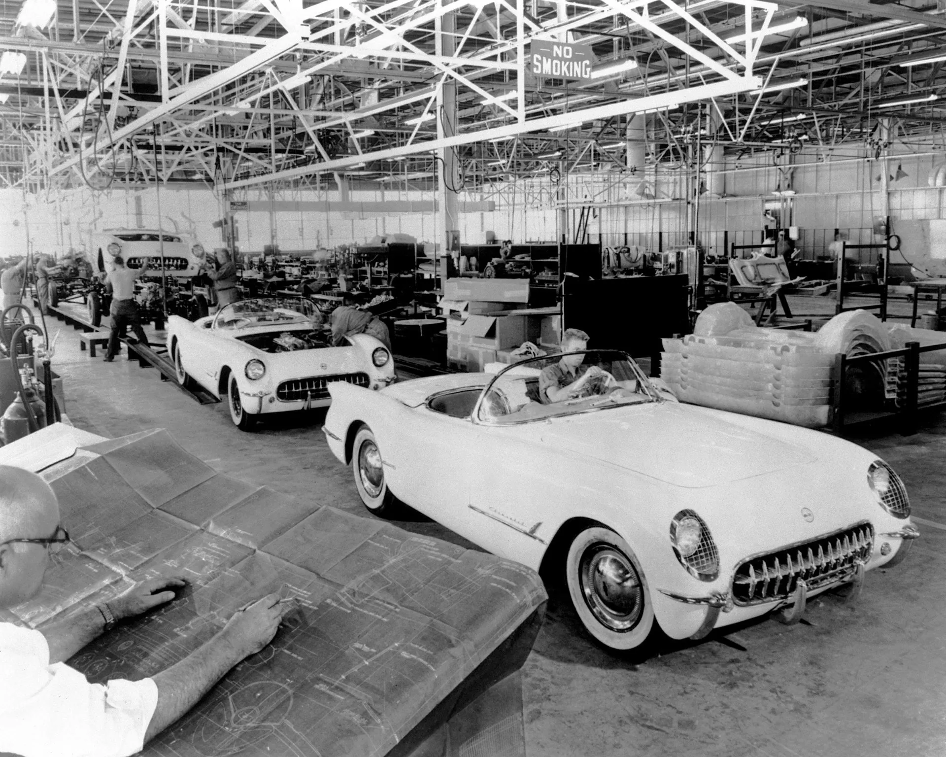 A black-and-white factory photo of white Corvette roadsters on an assembly line under steel roof trusses, a worker at the wheel of the nearest car, another car with its hood open behind it, stacked body parts at the right and a man reading blueprints in the foreground.