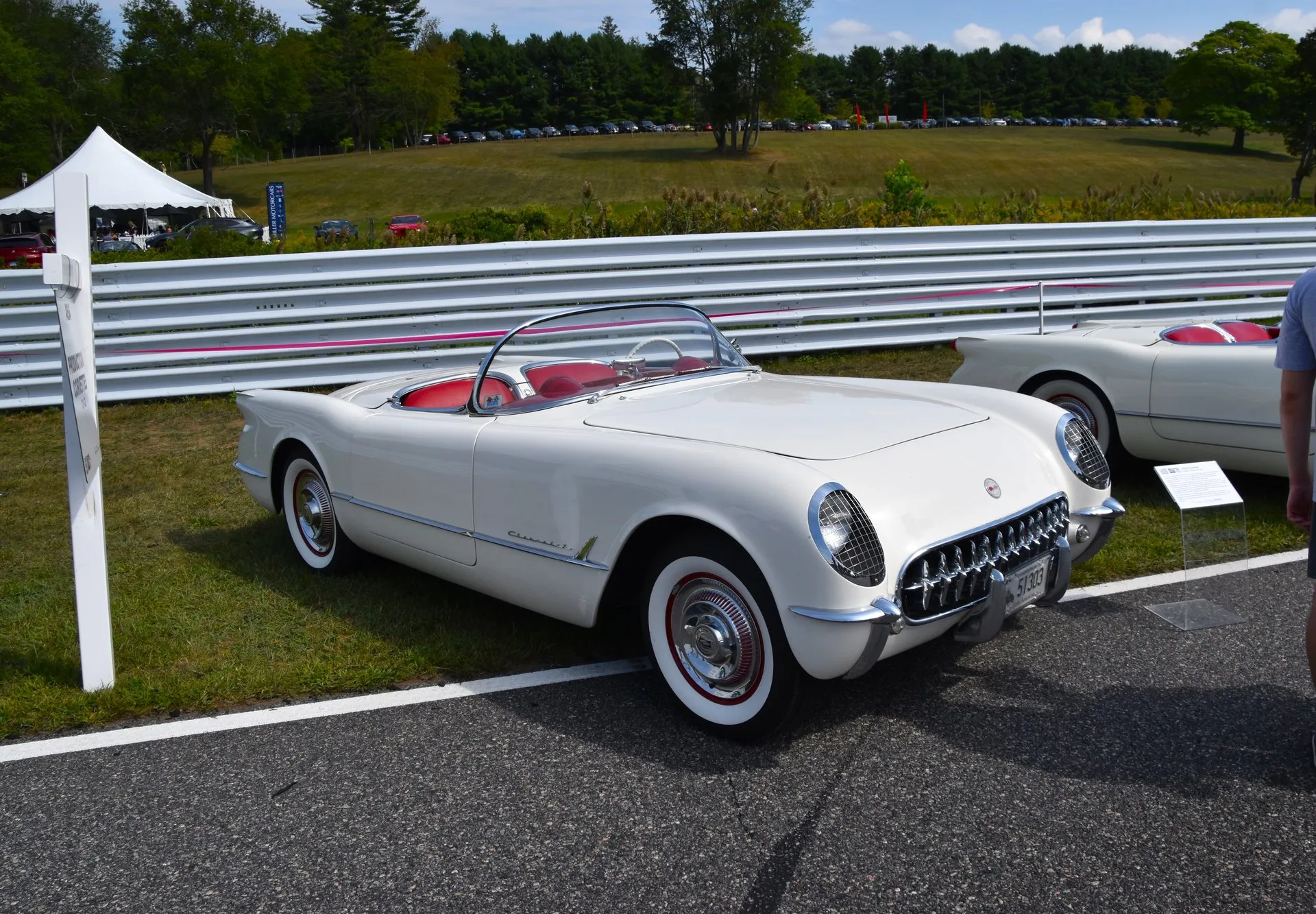 A white 1953 Corvette roadster with its top down and a red interior, parked on grass beside a racetrack barrier, seen from the front right, with whitewall tires, red wheels and chrome wheel covers, and another white early Corvette at the right edge.