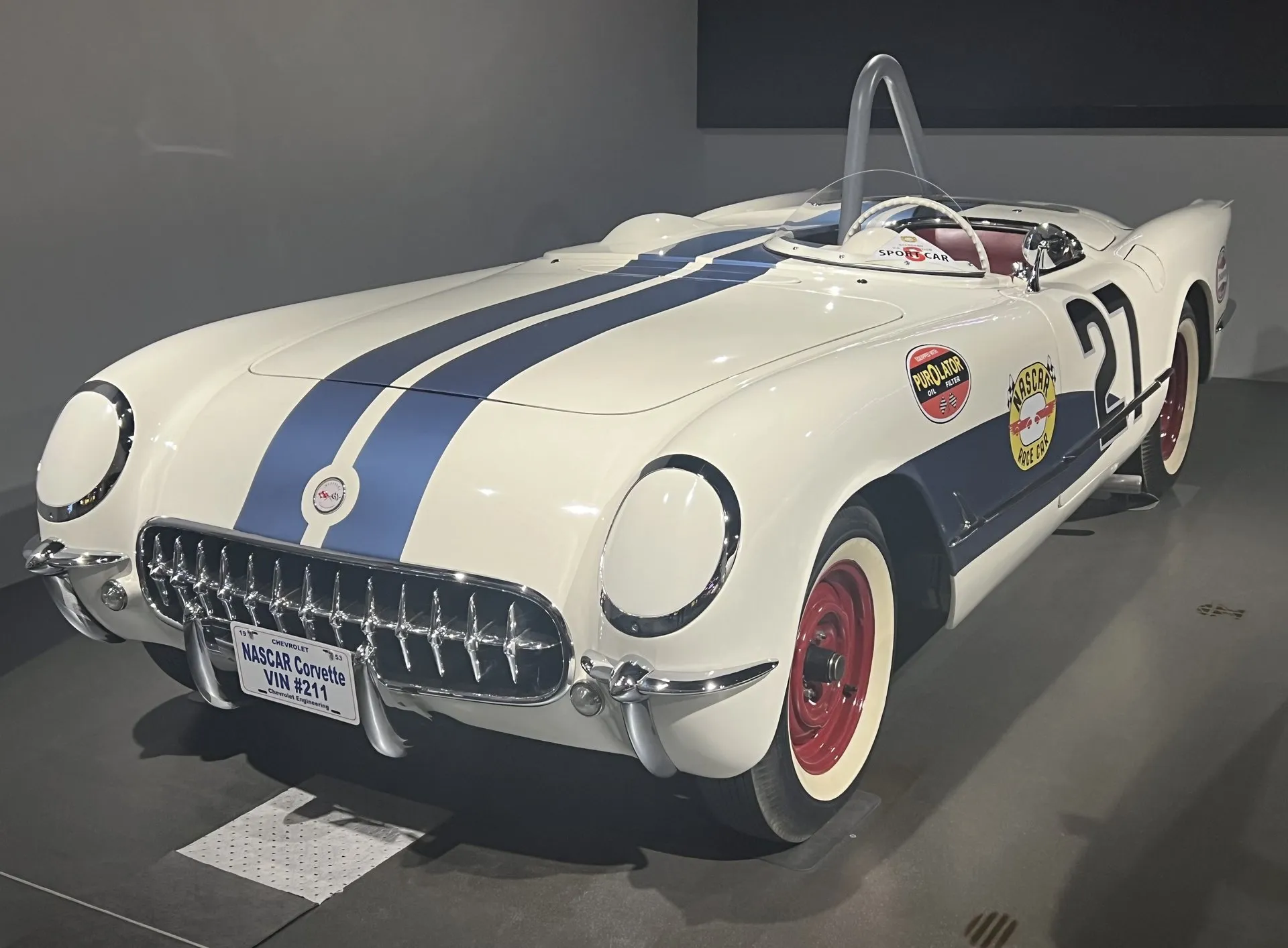 A white 1953 Corvette race car in a museum with twin blue stripes over the hood, a blue side panel, race number 27, red wheels, a roll hoop behind the seat and a front plate reading NASCAR Corvette VIN #211.