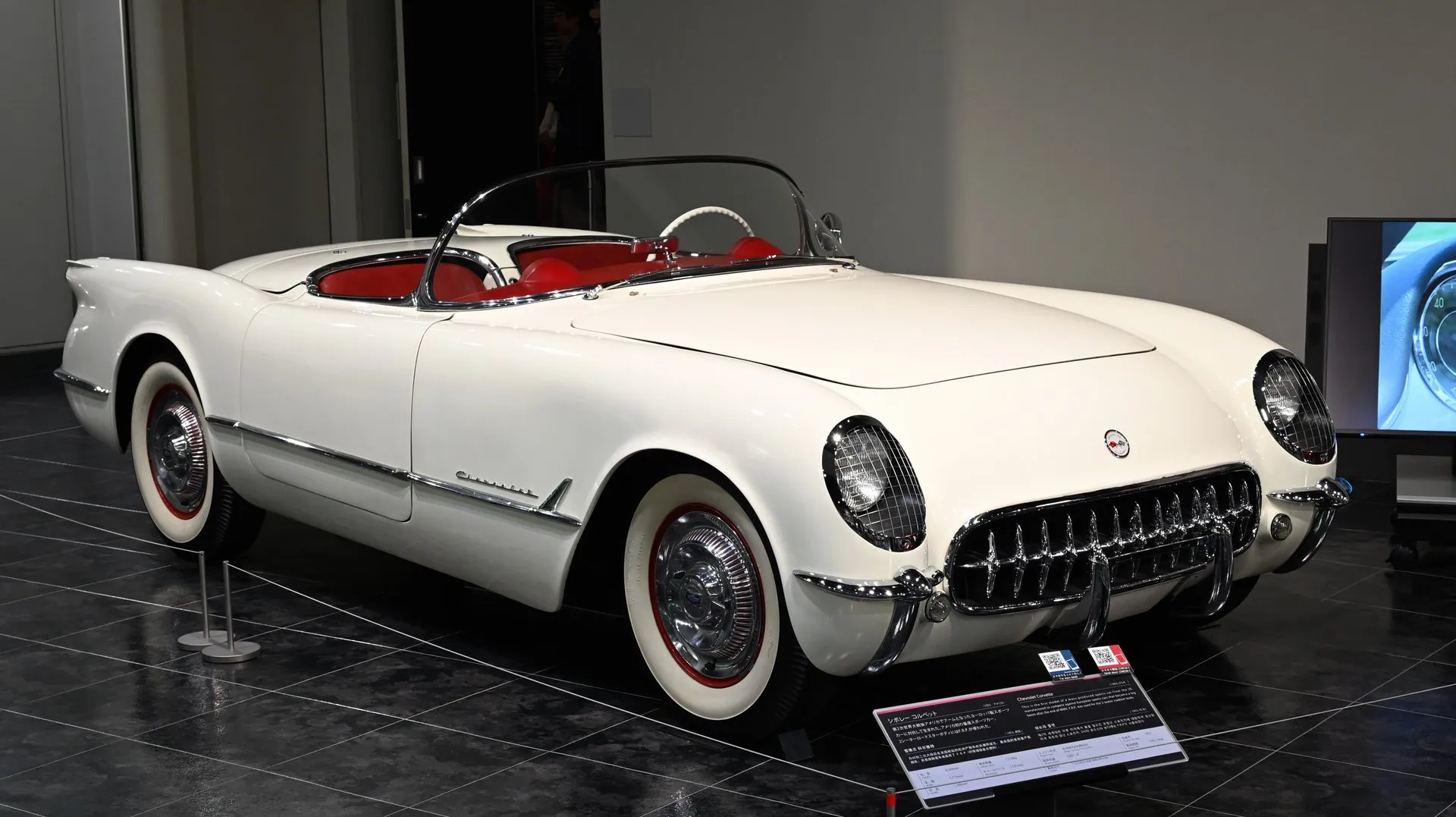 A white 1953 Corvette roadster with a red interior and whitewall tires on a dark museum floor, seen from the front corner, with chrome mesh guards over the headlights and a toothed chrome grille.