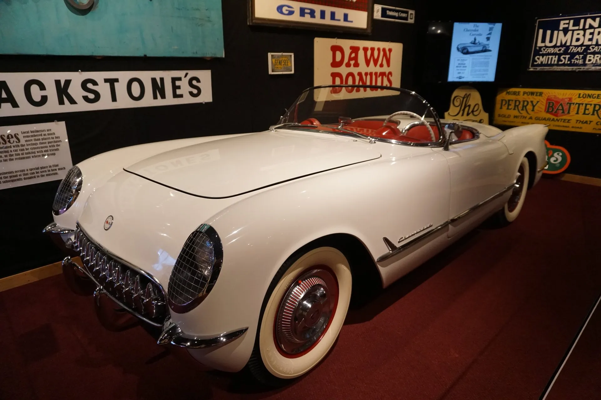 A white 1953 Corvette roadster with its top down and a red interior, seen from the front left in a museum, with mesh guards over the headlights, a toothed chrome grille, whitewall tires on red wheels with chrome covers, and old Flint shop signs on the wall behind.