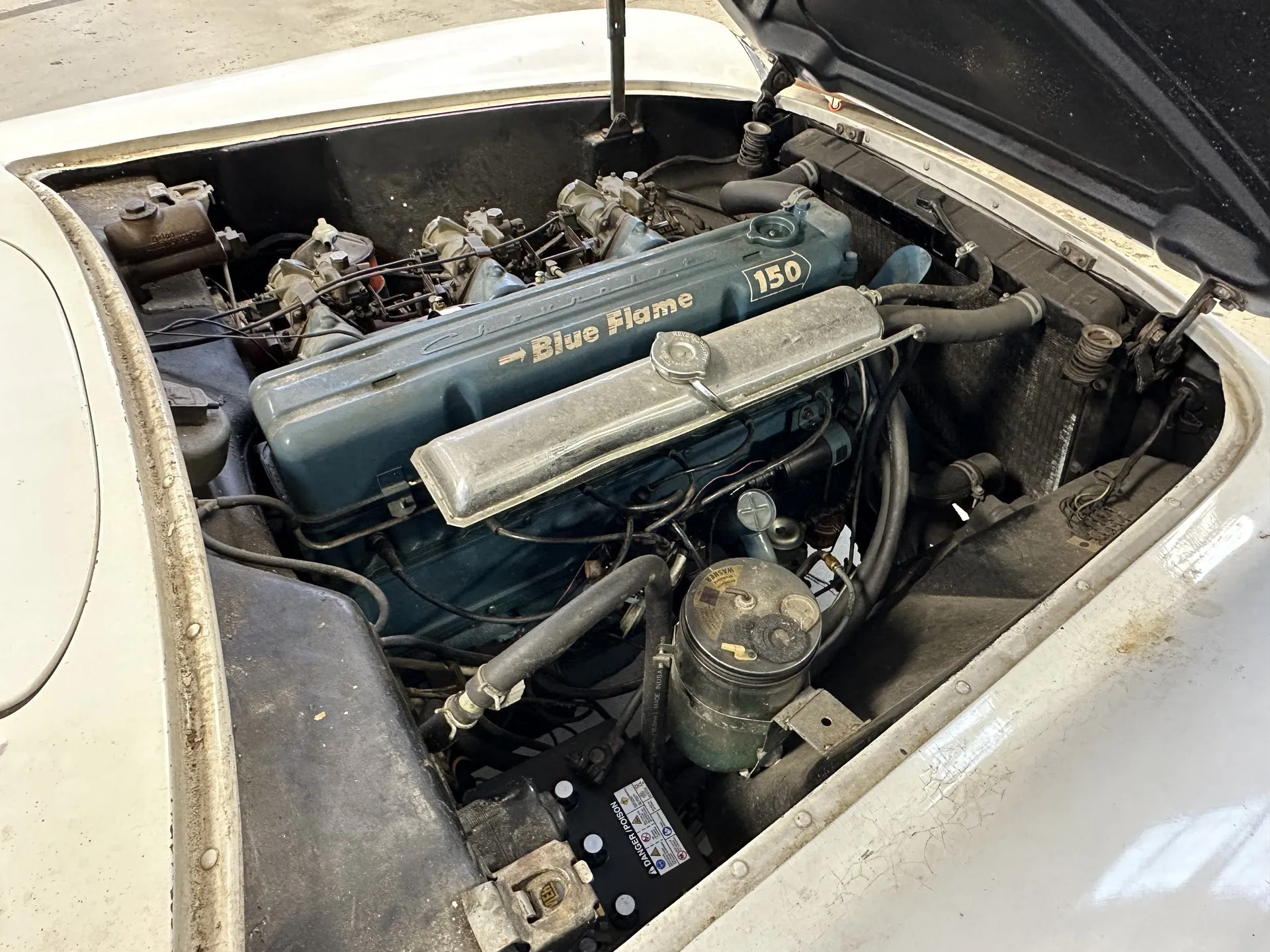 The engine bay of a white early Corvette with the hood propped open: a blue-gray inline six whose valve cover carries Blue Flame and 150 decals, three carburetors along its left side and a chrome cover over the spark plug wiring.