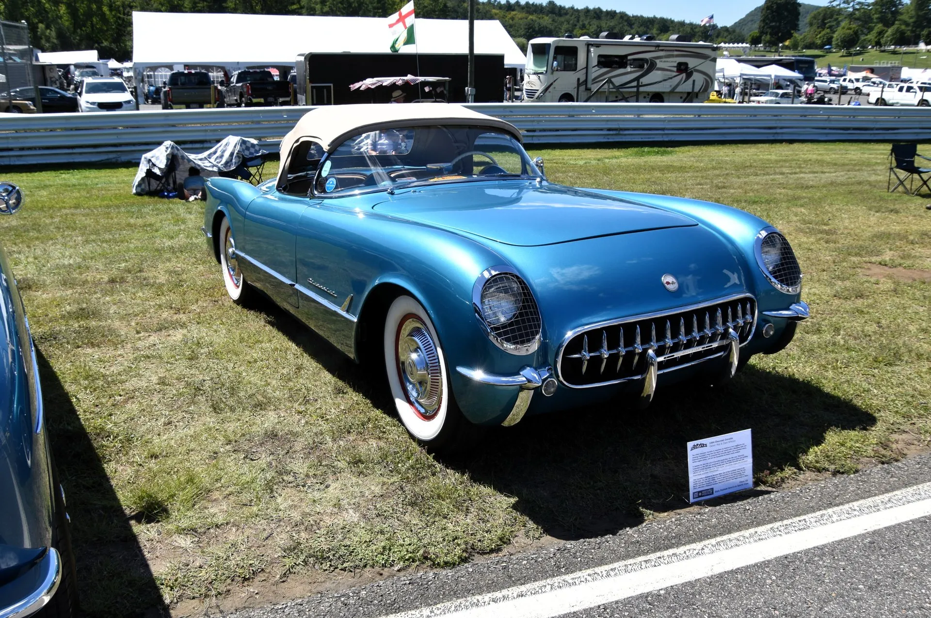 A metallic blue early Corvette roadster with a beige soft top up, parked on grass beside a racetrack, seen from the front right, with mesh guards over the headlights, a toothed chrome grille and wide whitewall tires.