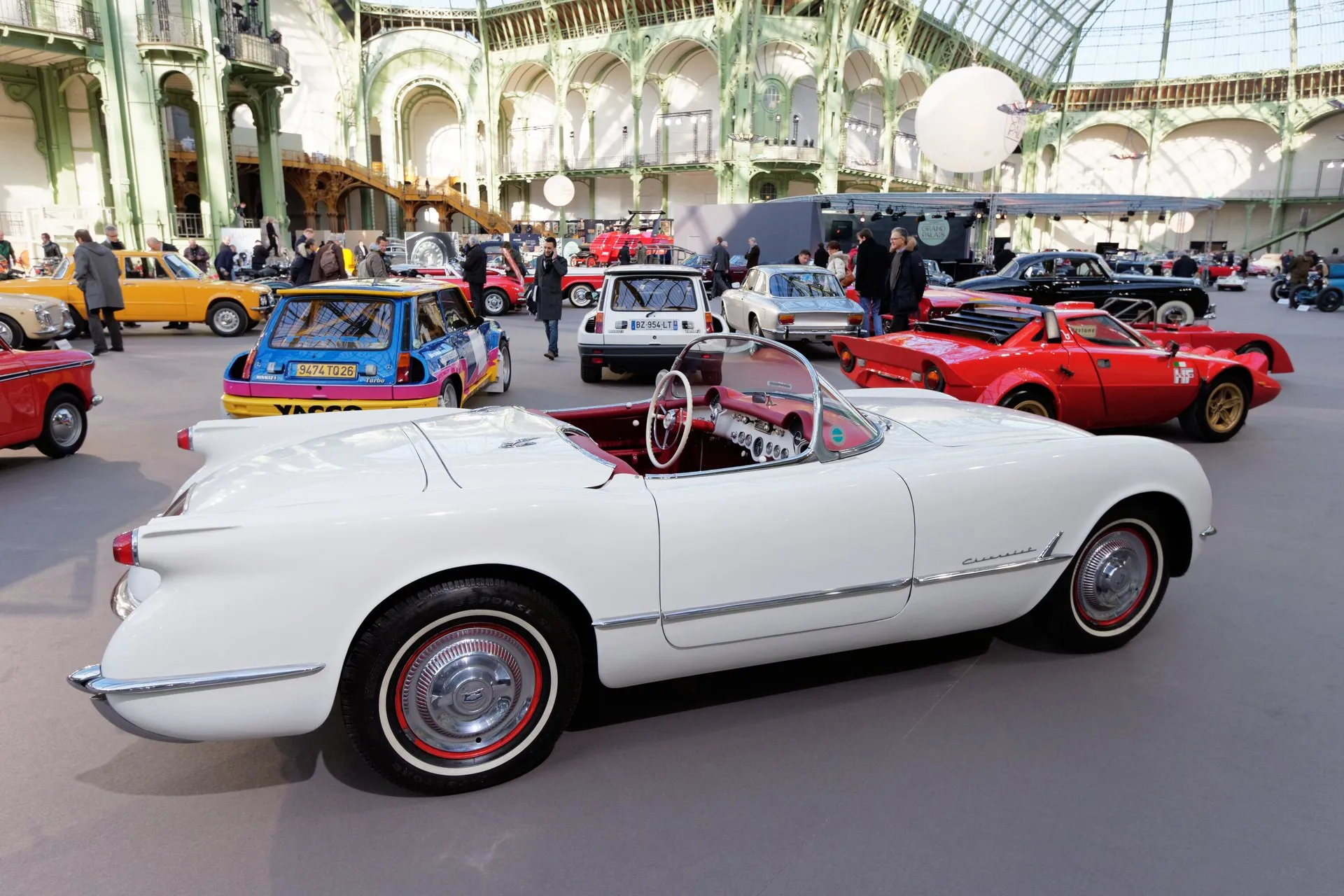 A white early Corvette roadster with its top down, red interior and whitewall tires on red wheels, seen from the right side on an auction floor under a glass and iron roof.