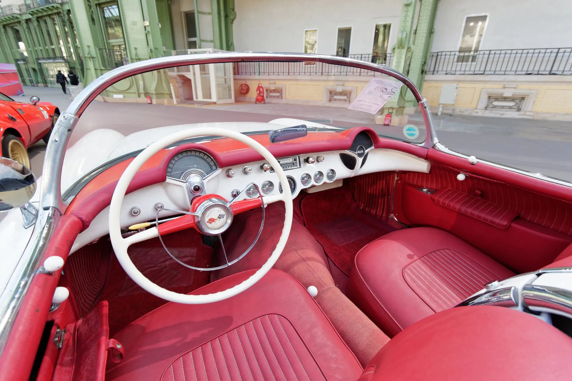 The cockpit of a white early Corvette: red seats, door panels and carpet, a white two-spoke steering wheel and a white instrument panel with a red padded crown and a row of round gauges.