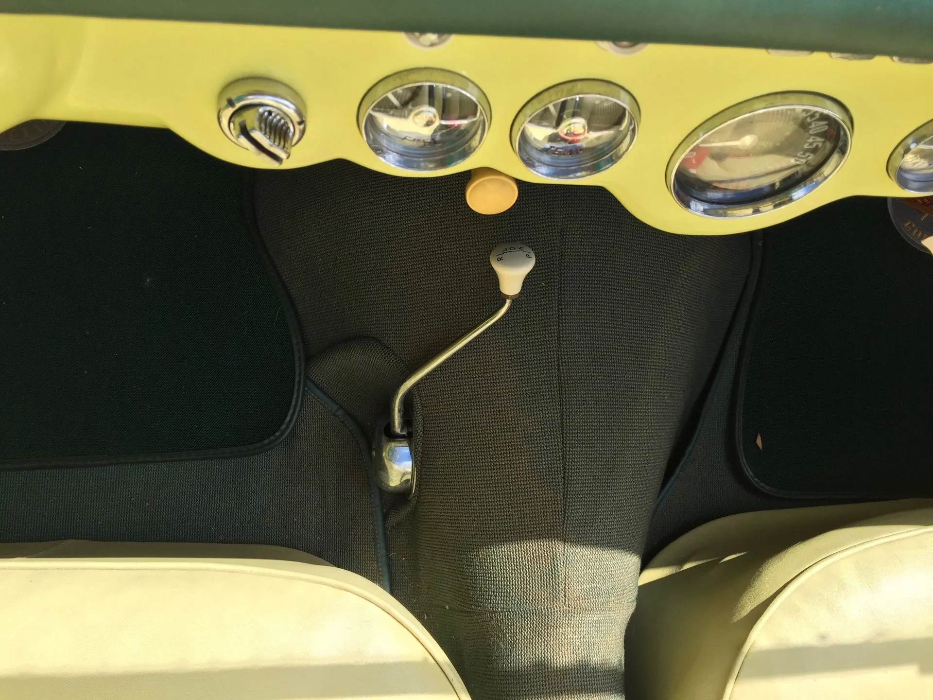 Looking down at the floor of a C1 Corvette between the seats: a chrome lever with a white knob rising from the transmission tunnel, under a pale yellow dash with round gauges.