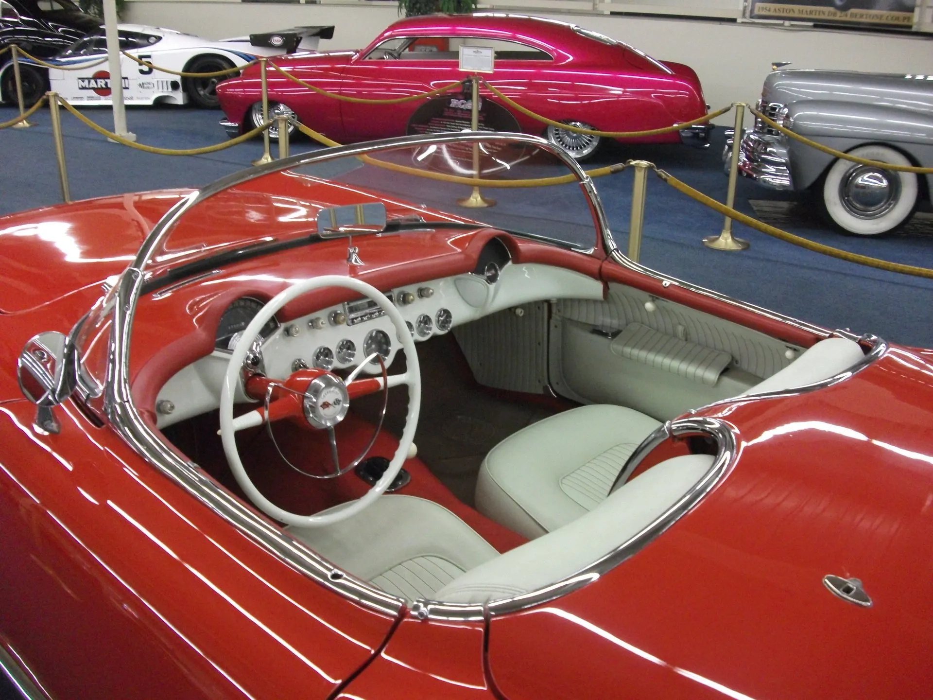 The open cockpit of a red C1 Corvette seen from above the driver's door: a white steering wheel, a row of round gauges in a white dash, white seats and a chrome windshield frame, with no side windows fitted.