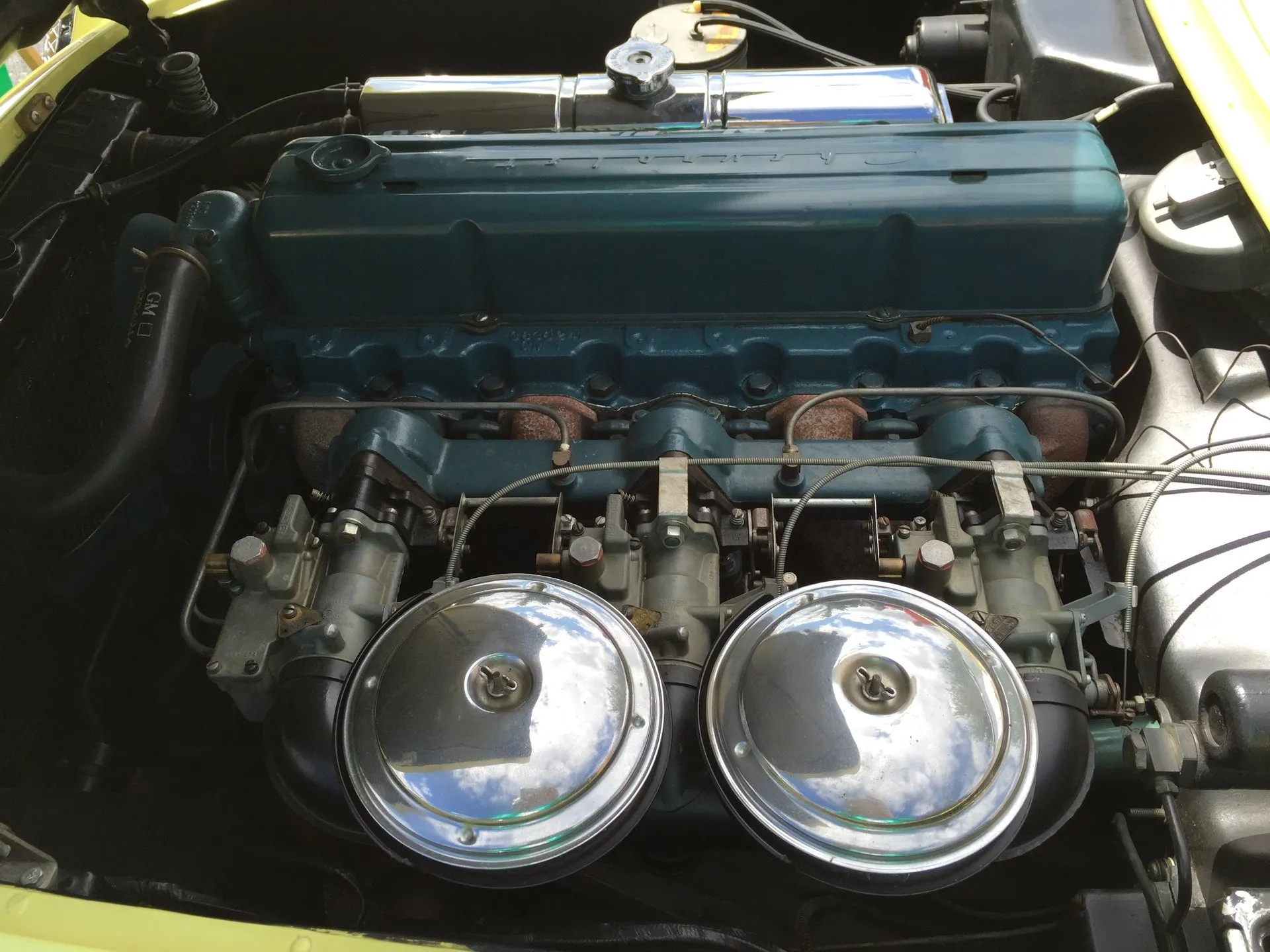 A blue inline six-cylinder engine in a C1 Corvette engine bay, with three side-draft carburetors under two round chrome air cleaners.