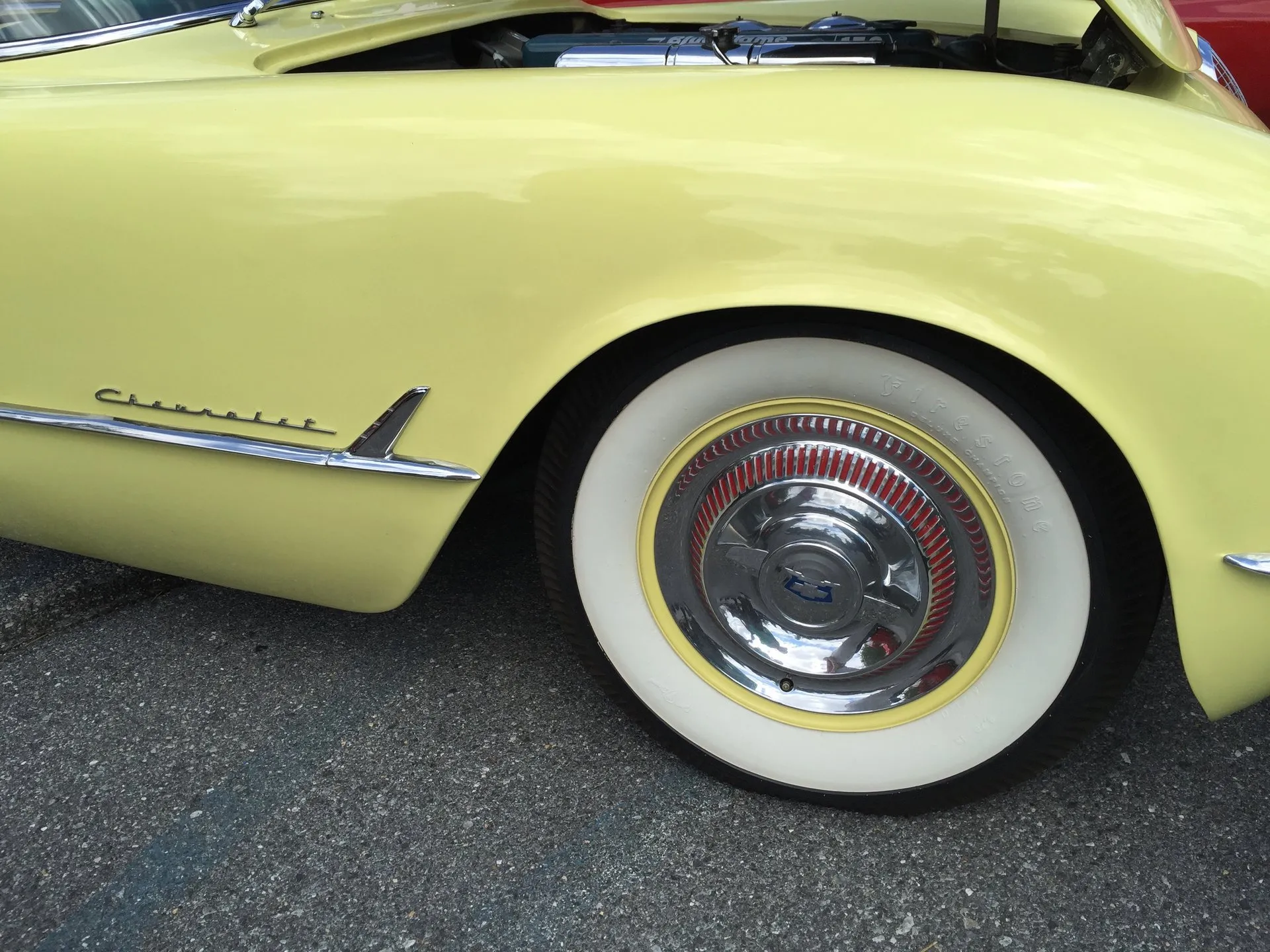 The front fender of a pale yellow C1 Corvette: a plain chrome Chevrolet script with no gold V, a whitewall tire on a yellow wheel with a chrome spinner cover, and the open hood showing a blue valve cover.