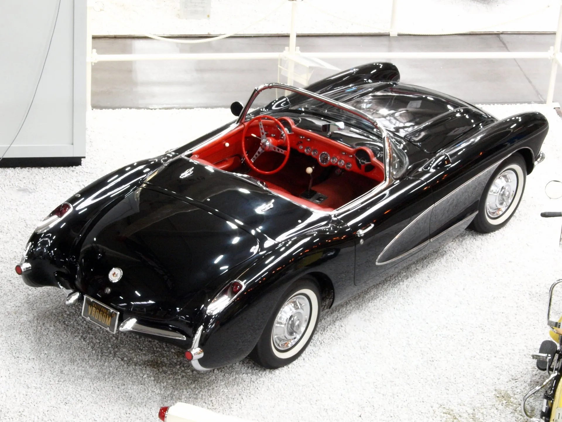 A black C1 Corvette convertible with a red interior, seen from above and behind in a museum hall: a red dash with round gauges, a red steering wheel, a floor shifter with a white knob, chrome-edged side coves and whitewall tires.