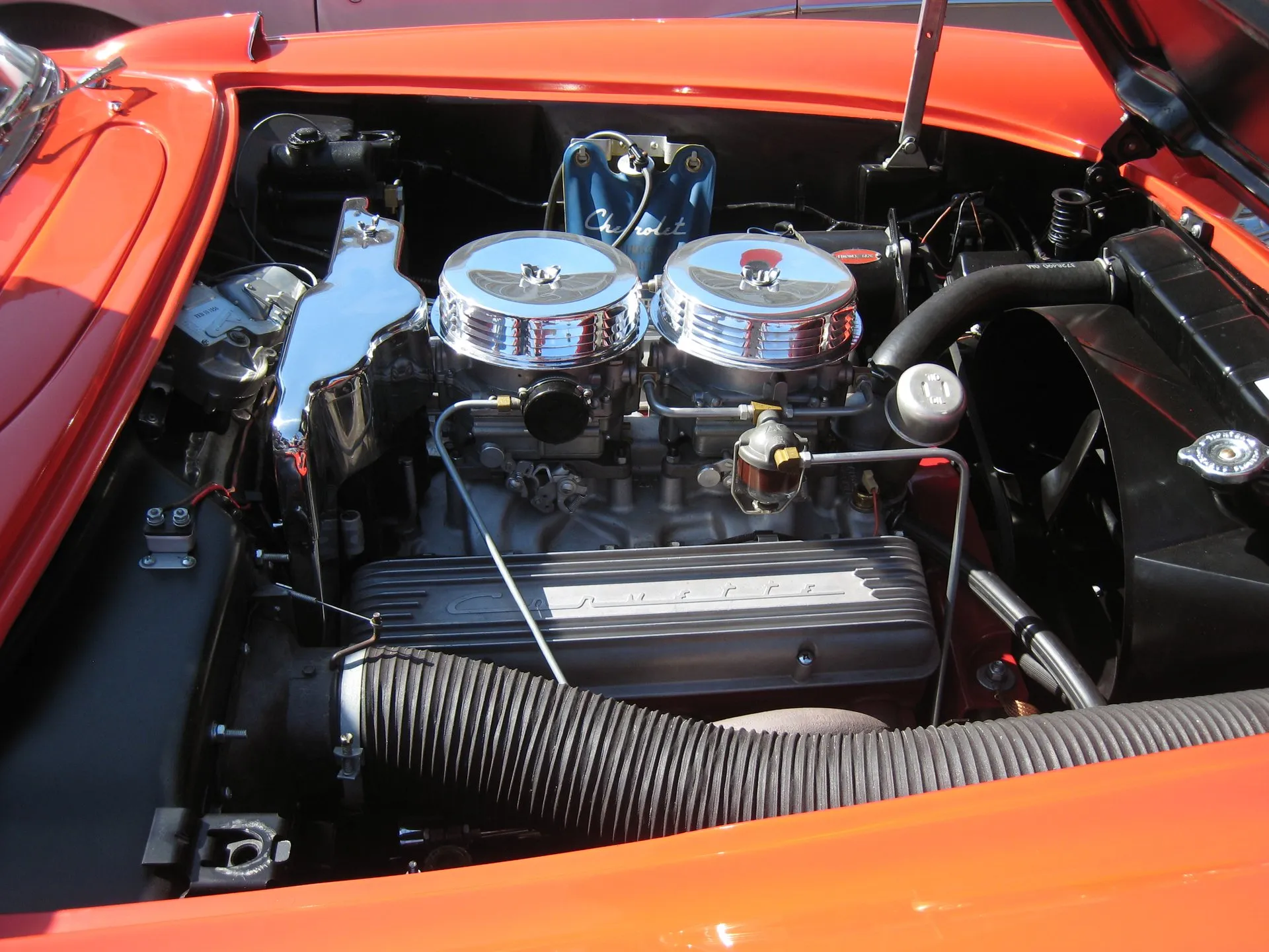 The engine bay of a red C1 Corvette: two four-barrel carburetors under round chrome air cleaners, a finned valve cover with Corvette script, a blue vinyl washer bag on the firewall and a large ribbed hose.