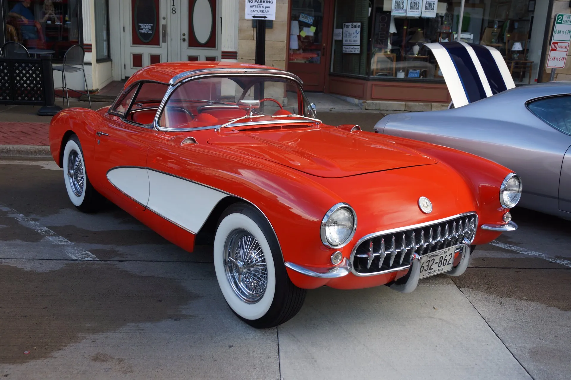 A red C1 Corvette with white side coves, whitewall tires, wire wheels and a red removable hardtop, parked at a curb in front of shops, seen from the front left.