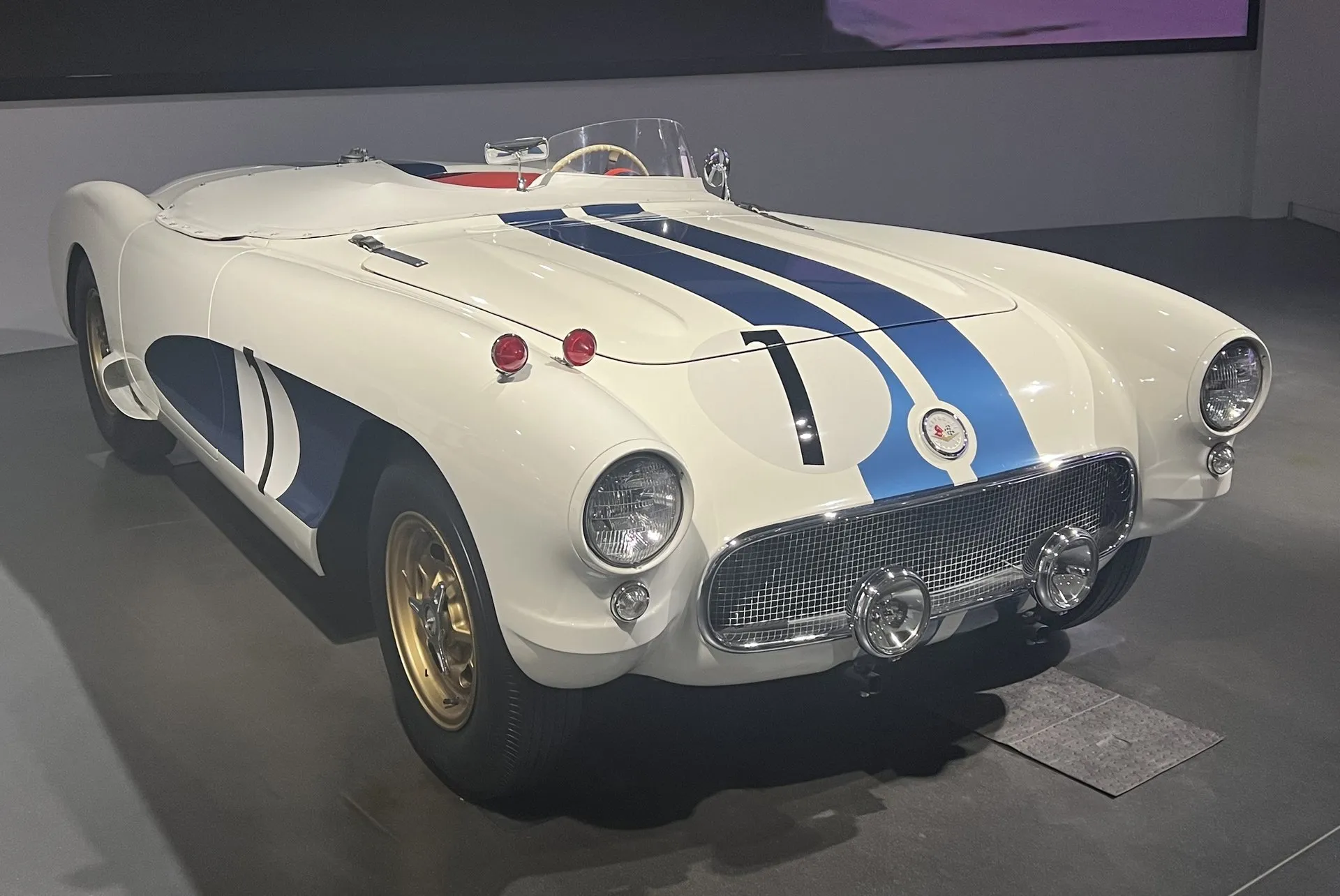 A white C1-based race car with two blue stripes over the hood, a blue side cove carrying the number 1, a small racing windscreen, gold wheels and two driving lamps, on display in a museum.