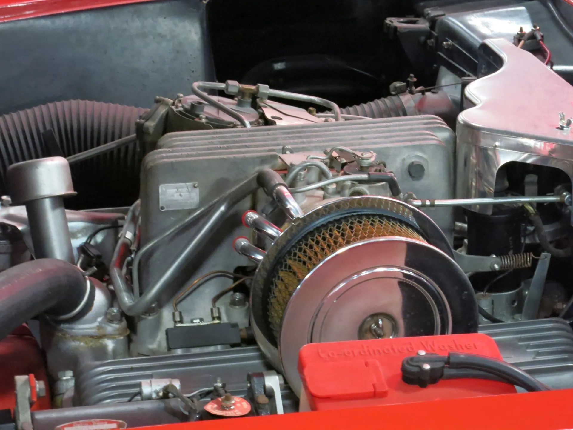 Close view of a fuel-injected V8 in a red C1 Corvette: a finned aluminum injection plenum with a small data plate, a round chrome air cleaner over a mesh-covered filter, chrome tubes, a finned valve cover and a red Co-ordinated Washer reservoir in front.