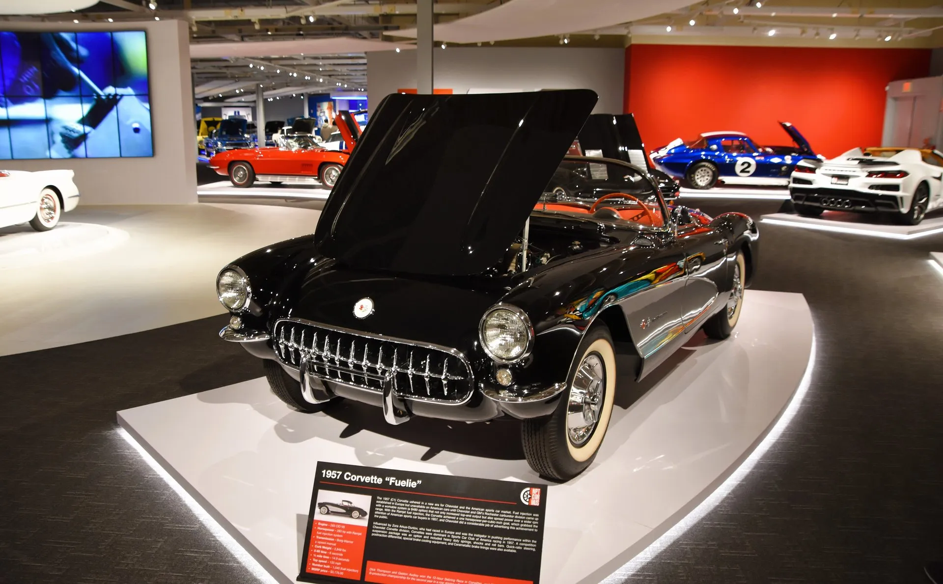 A black 1957 Corvette with whitewall tires and a red interior on a lit museum stand, hood raised, seen from the front left, behind a placard titled 1957 Corvette Fuelie.