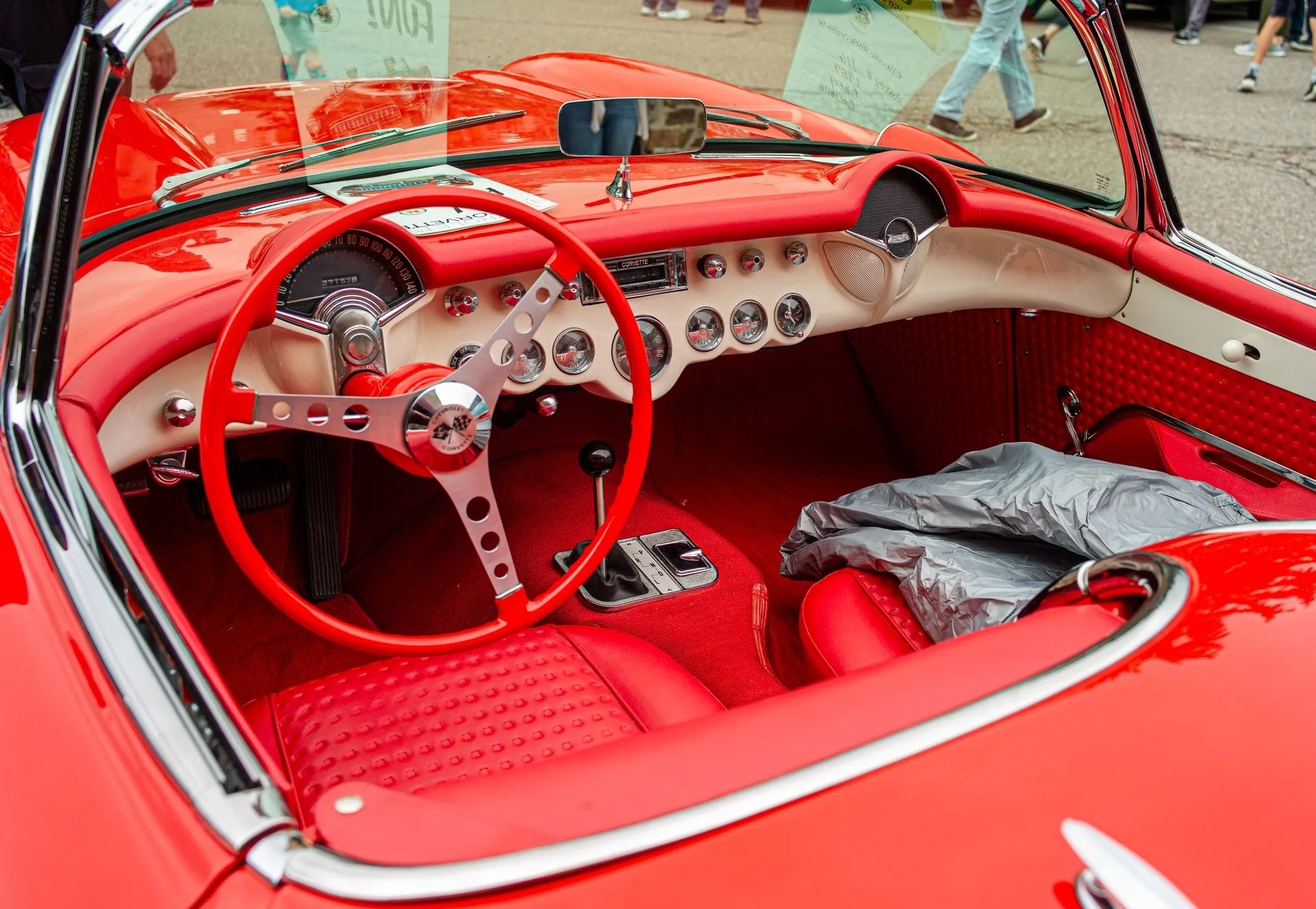 The red interior of a C1 Corvette seen from above the driver's door: a red three-spoke steering wheel, a speedometer in front of the driver, round gauges across a white dash with a large one in the middle, a floor lever with a P R N D L plate, red waffle-pattern seats and red carpet.