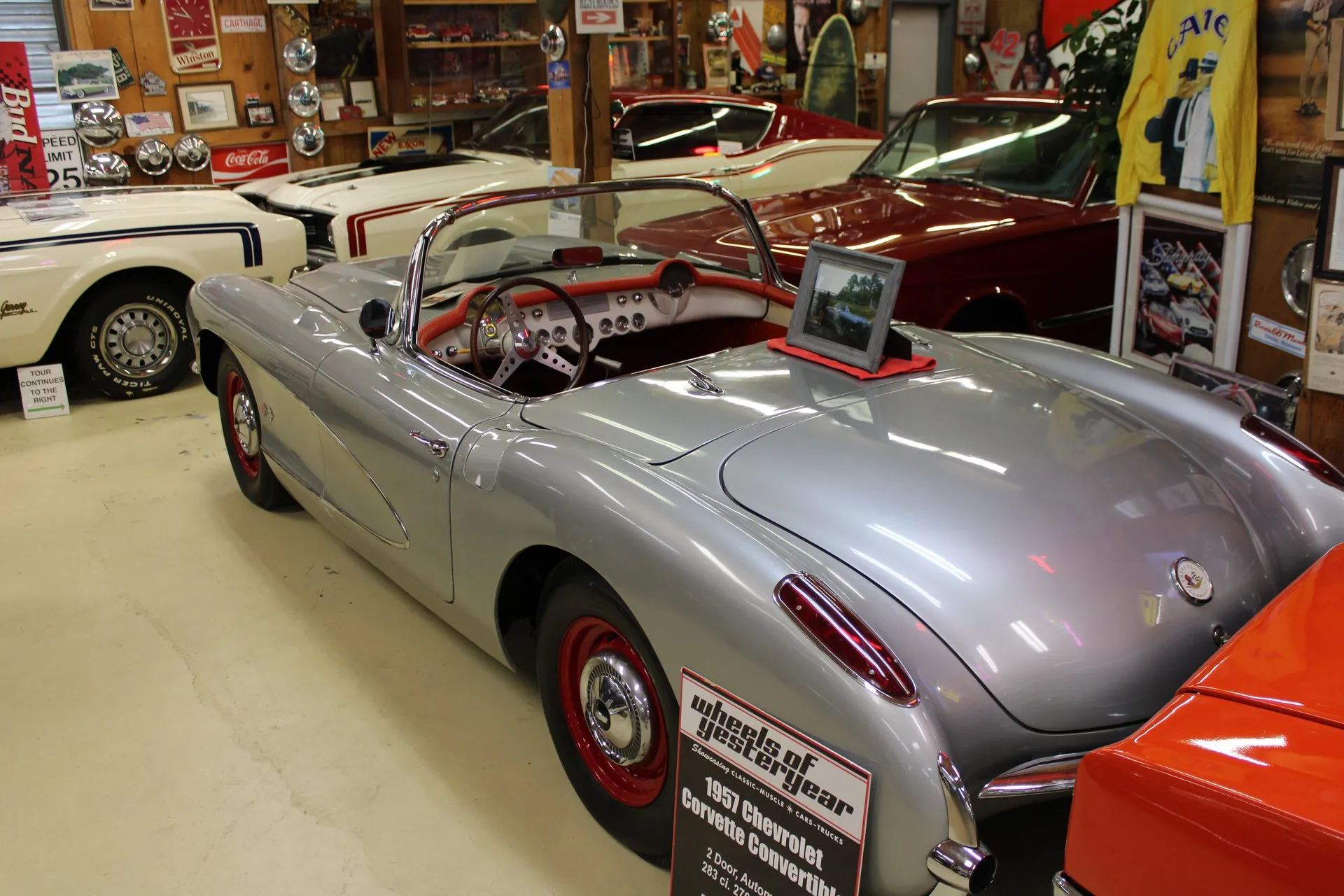A silver C1 Corvette convertible with its top down, a red interior and red-painted wheels with small chrome hubcaps, seen from the rear left in a museum hall full of cars and signs, with a placard reading 1957 Chevrolet Corvette Convertible.