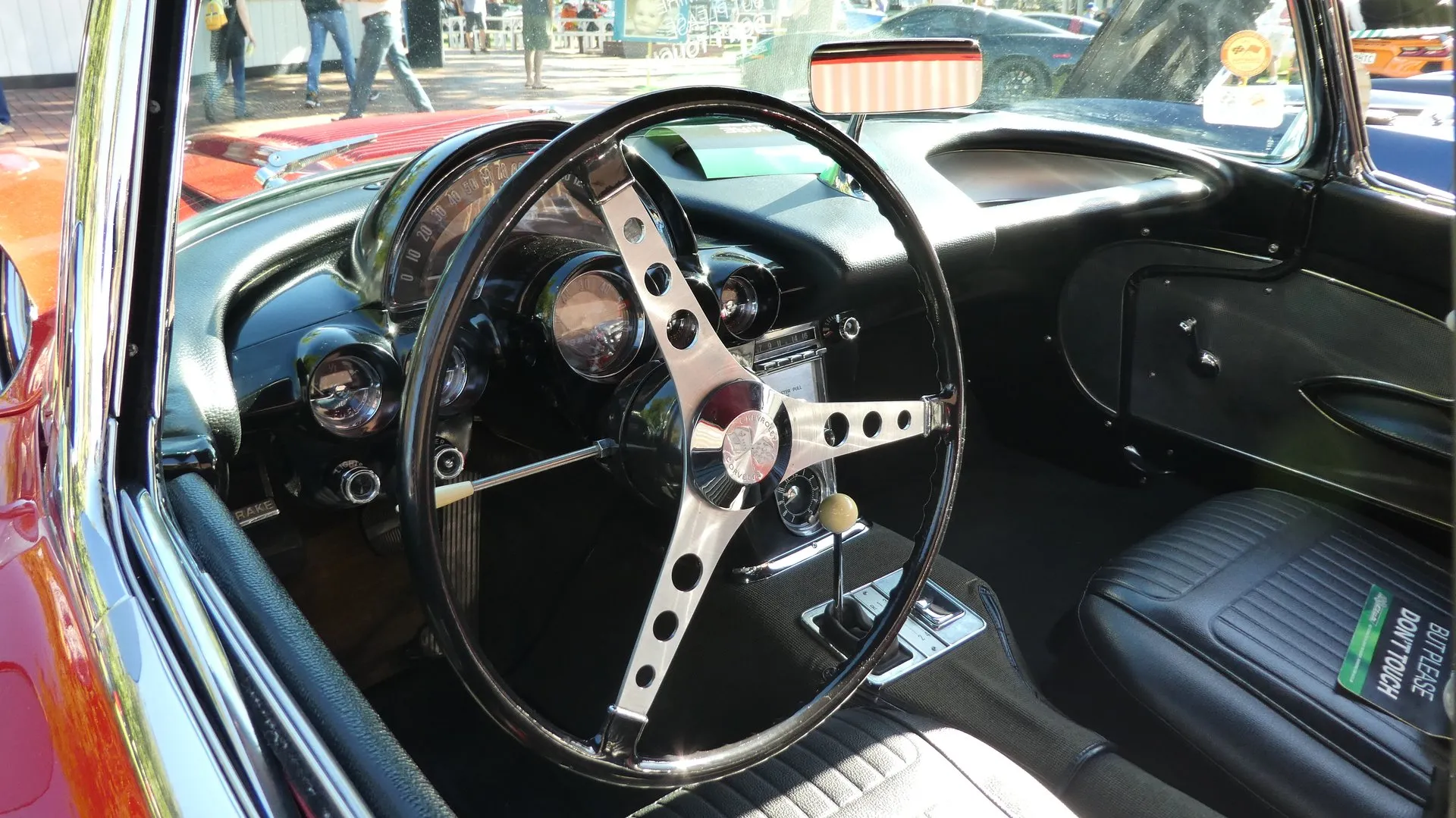 The cockpit of a red C1 Corvette seen through the open driver's door: a three-spoke steering wheel, a speedometer arc above the column with a round tachometer under it, smaller gauges on either side, a center console with a radio and a floor shift lever, and dark vinyl seats.