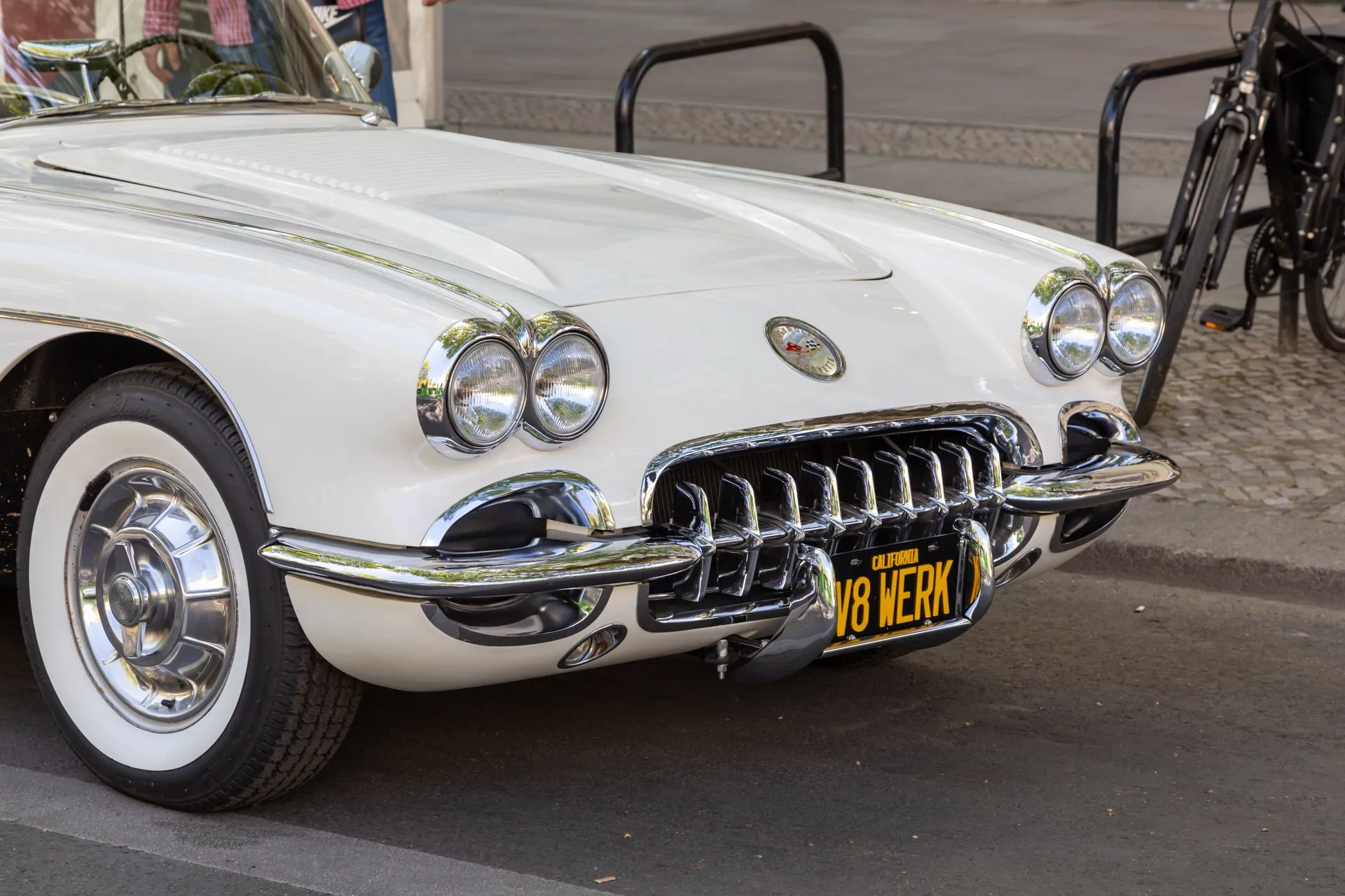 The front of a white 1958 Corvette: four headlights in chrome bezels, a chrome grille with vertical teeth, a round emblem on the nose and a ribbed louver panel on the hood, with a black-and-yellow California plate.