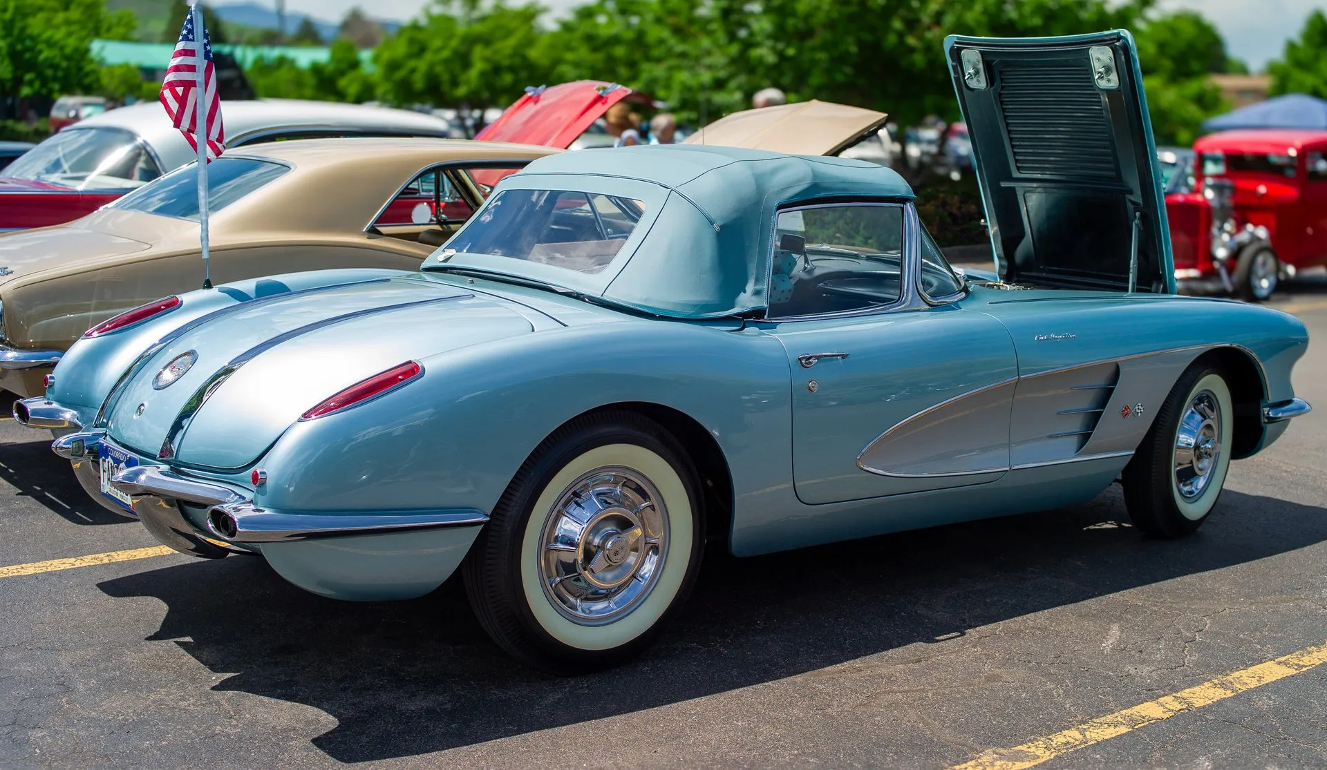 A light metallic blue 1958 Corvette with silver side coves and a blue-gray soft top, seen from the rear three-quarter in a car-show parking lot, with its hood raised, twin chrome spears running down the trunk lid and Fuel Injection script on the front fender.