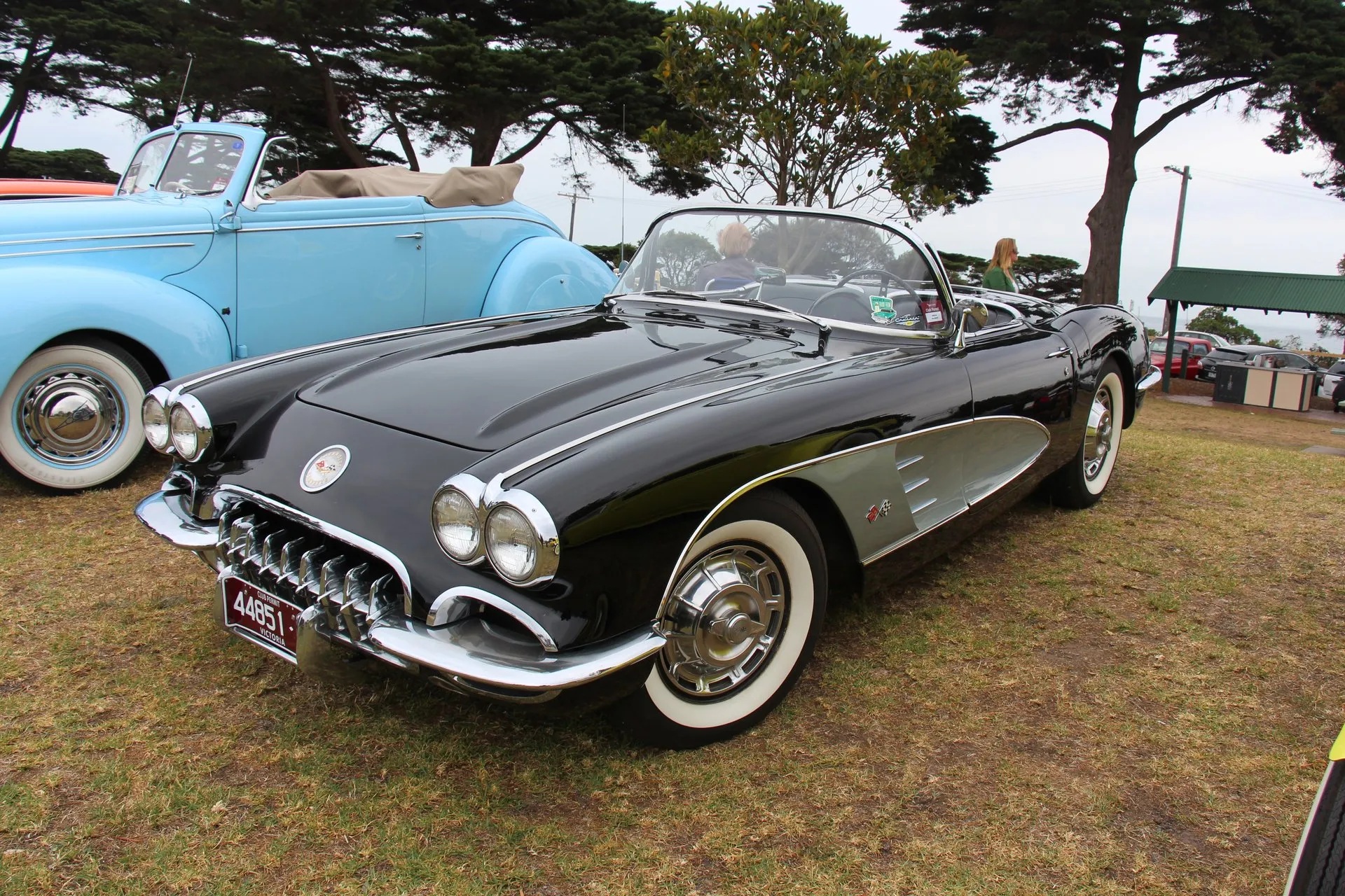 A black C1 Corvette with silver side coves and its top down, parked on grass at a car show, seen from the front right corner: four headlights, a toothed chrome grille, a smooth hood with no louvers, and whitewall tires with full wheel covers.