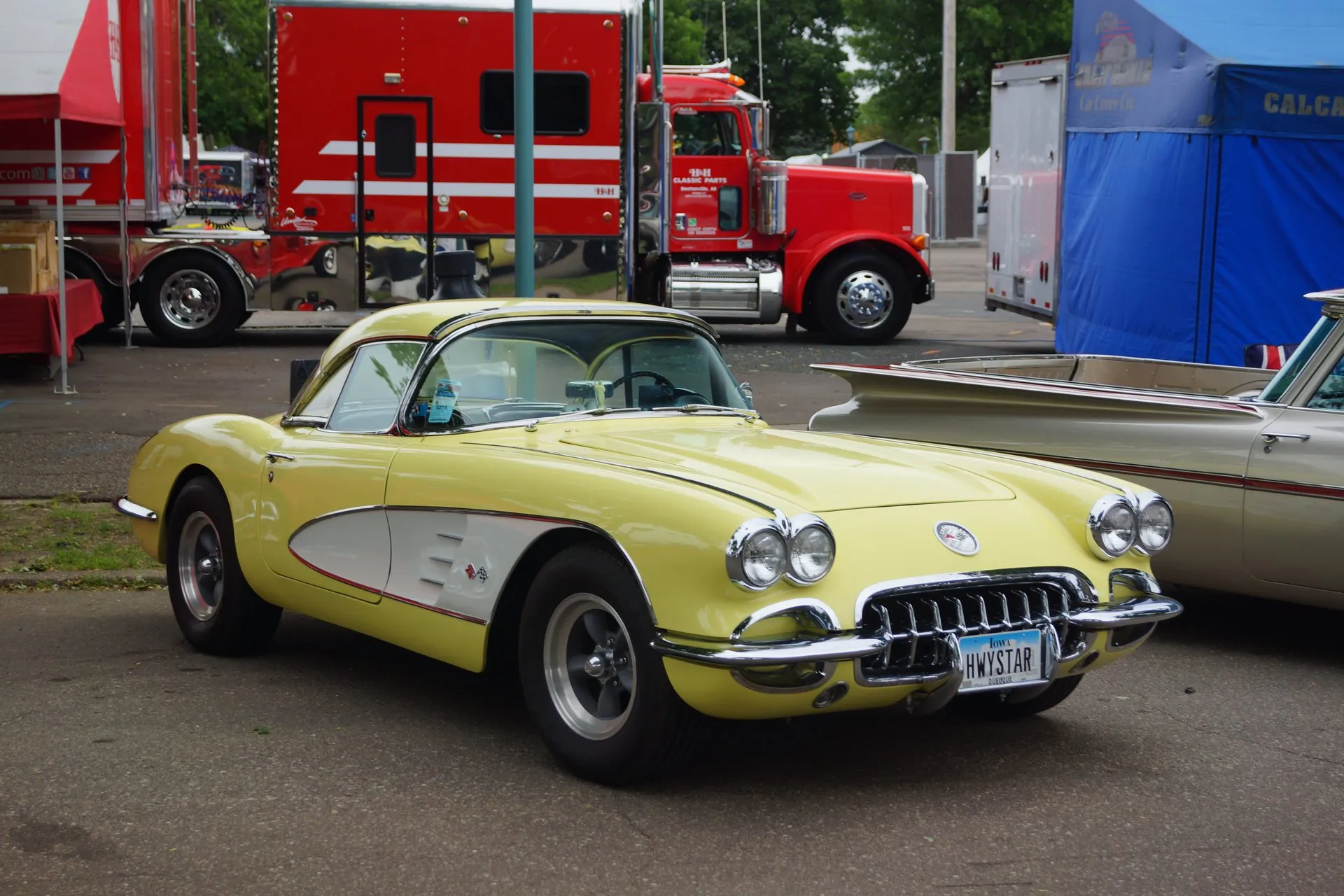 A pale cream C1 Corvette with a white side cove and a matching hardtop, parked on a lot in front of a red semi truck, wearing aftermarket slotted wheels and wide tires.