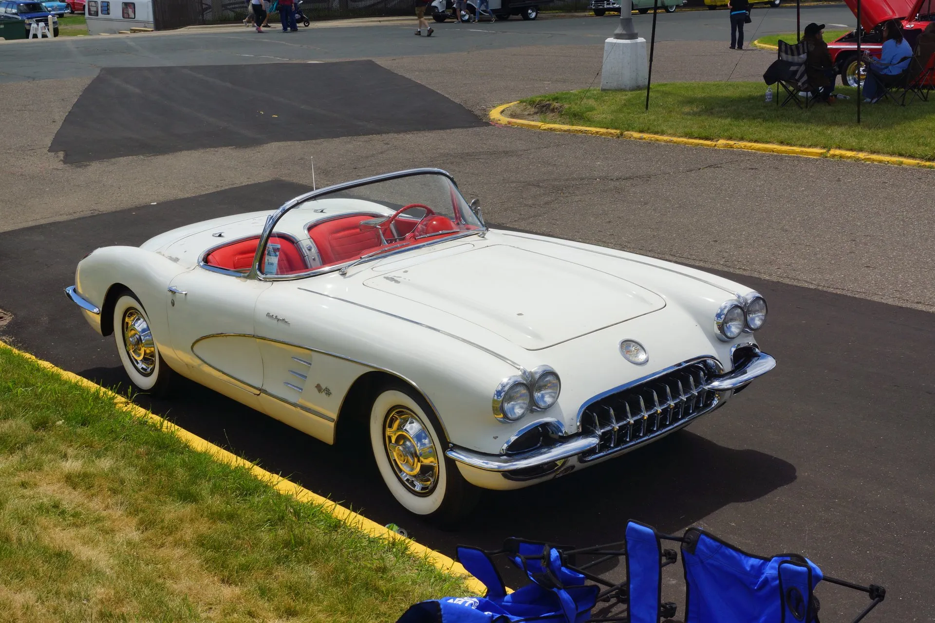 A white C1 Corvette with a red interior and its top down, seen from above and ahead on a car-show lot: a plain hood with two raised ridges and no louvers, a white side cove, whitewall tires and wheel covers with gold centers.