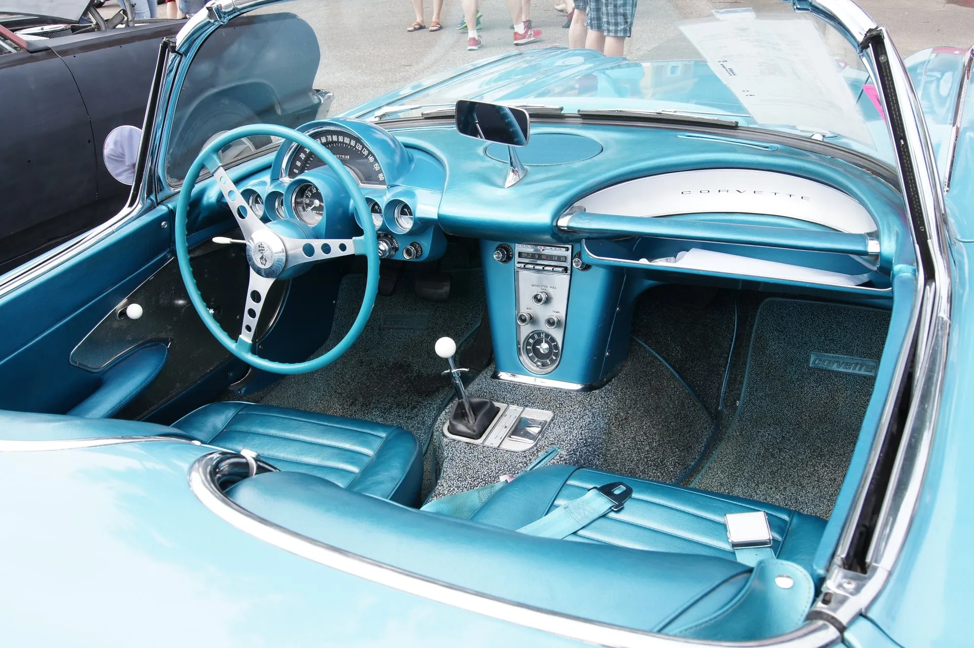 The turquoise cockpit of a C1 Corvette: a three-spoke steering wheel, the arched speedometer over the column with gauges below, a center console with radio and clock, a floor shifter with a white knob, a passenger grab bar with an open storage bin beneath it, and seats with pleats running side to side.
