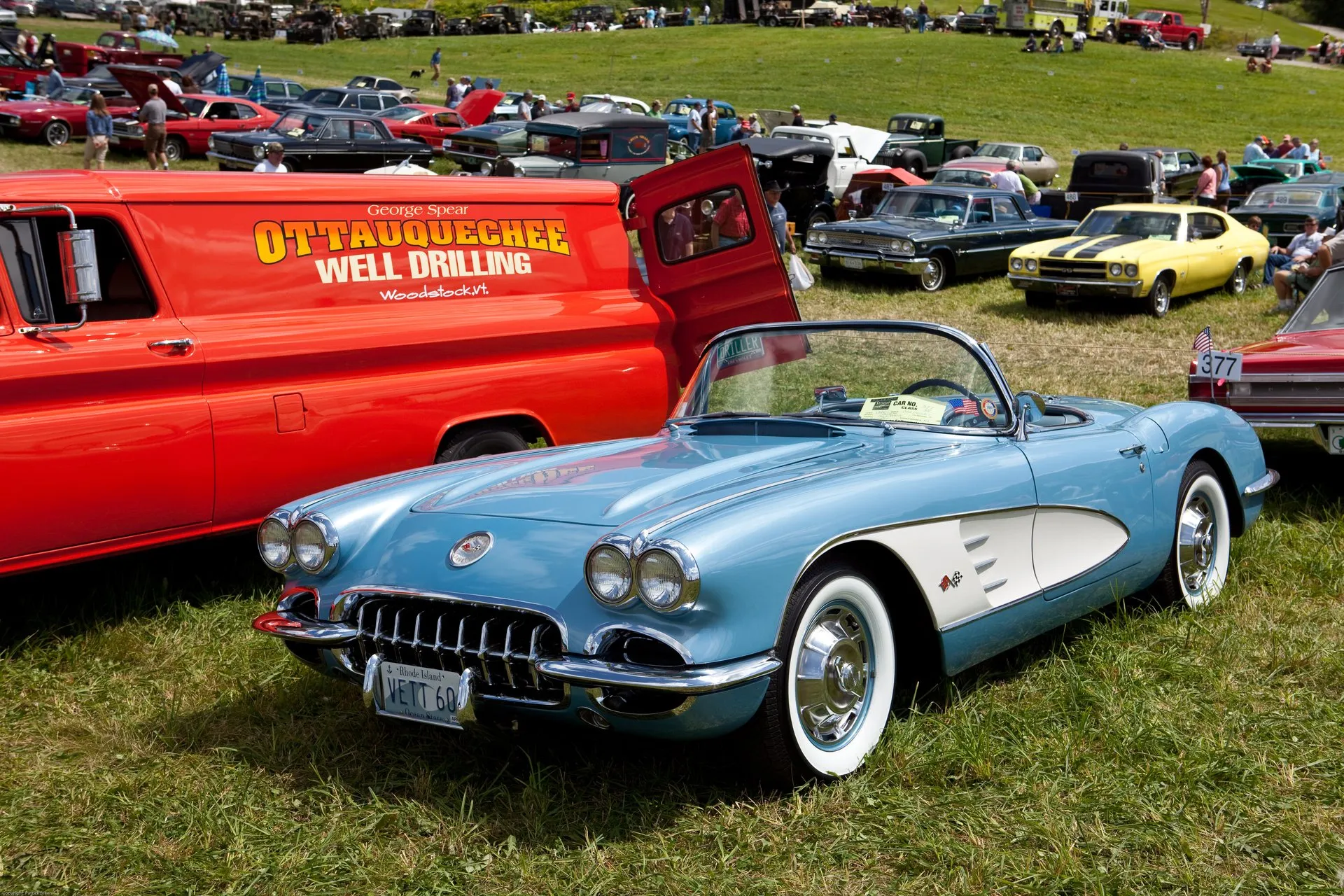 A light blue C1 Corvette with white side coves and its top down, parked on grass at an outdoor car show beside a red panel van, with whitewall tires, full wheel covers and a toothed chrome grille.