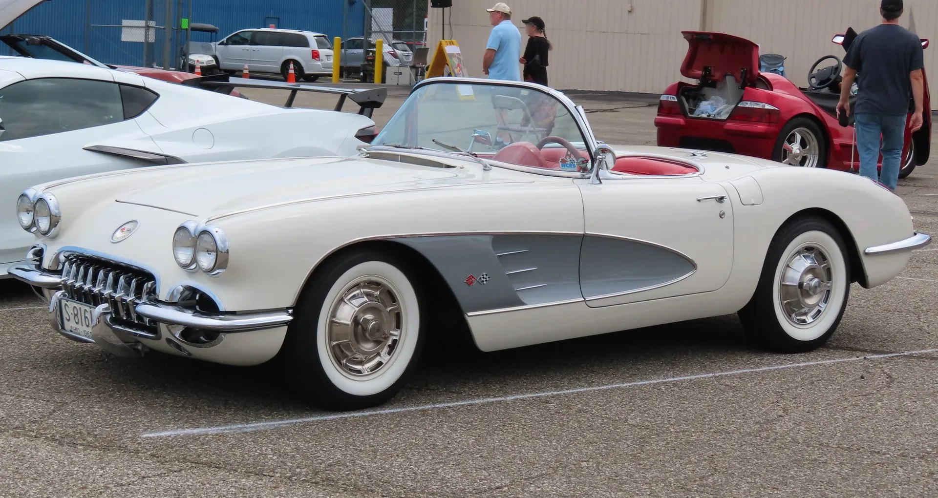 A white C1 Corvette with silver side coves, a red interior and its top down, parked on an airport apron at a car show, seen from the front left, with quad headlights, a toothed chrome grille, whitewall tires and full wheel covers.