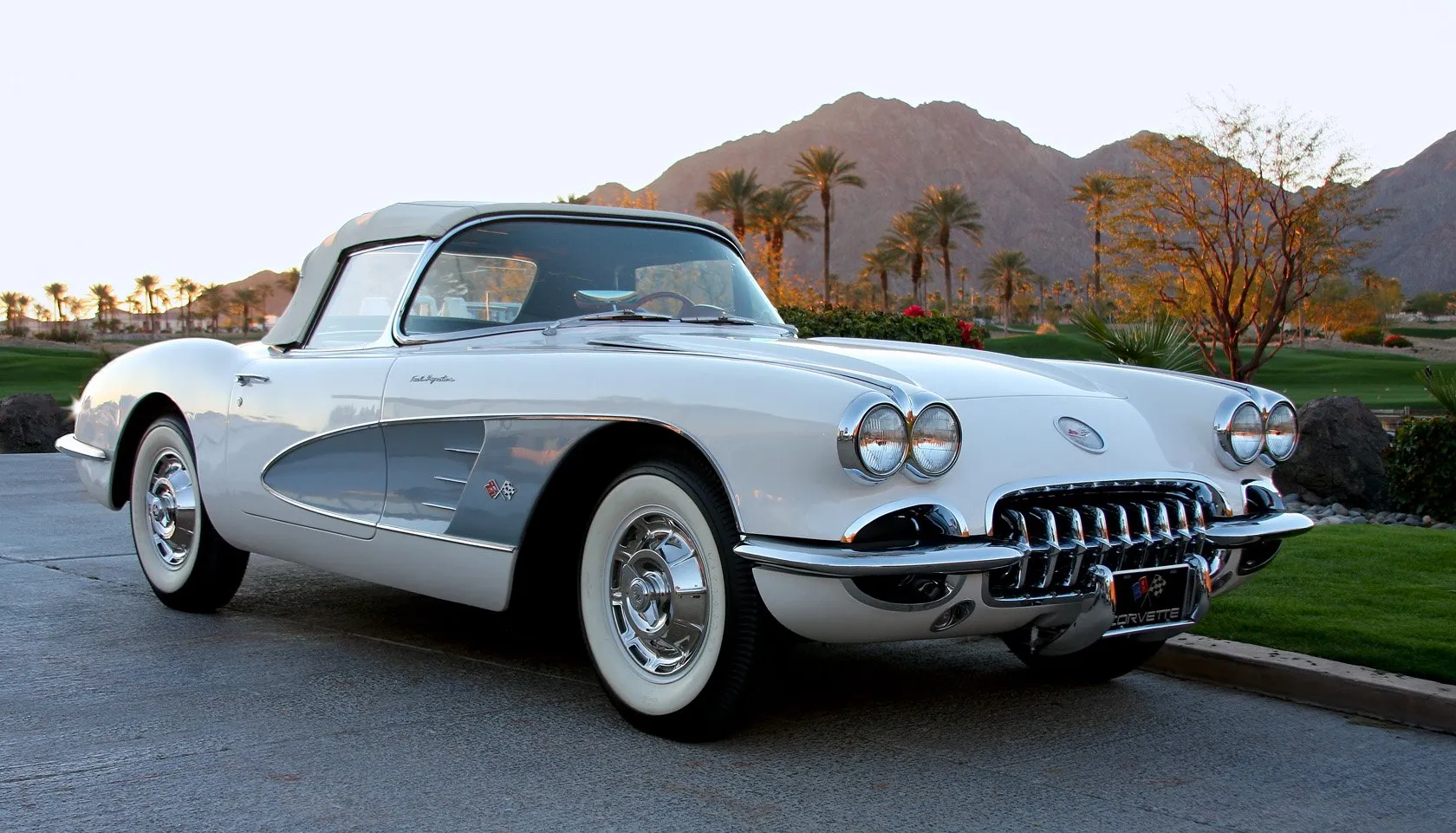 A white C1 Corvette with silver side coves and its top up, parked at dusk in front of palm trees and desert mountains, with small Fuel Injection script on the front fender and a toothed chrome grille.