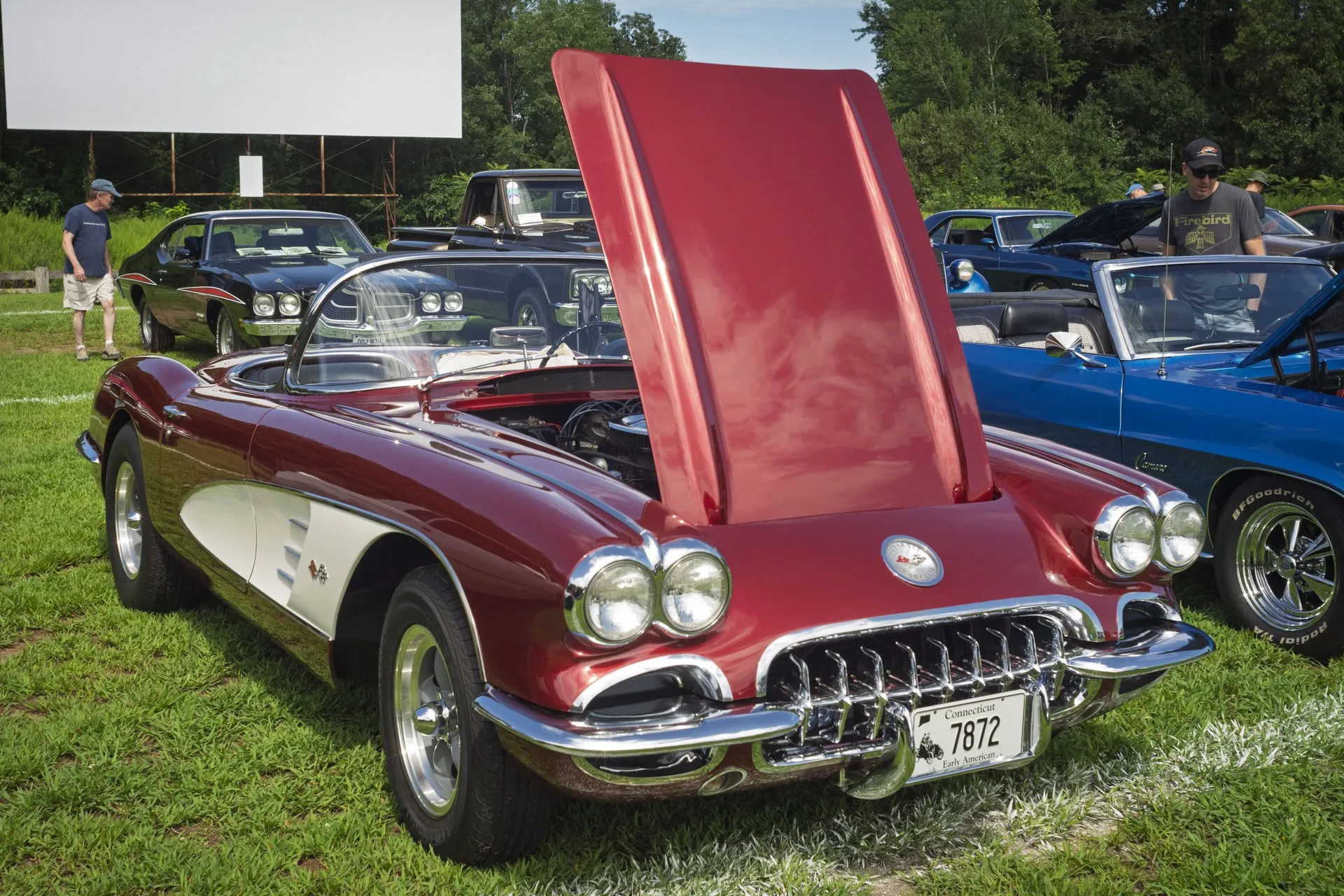 A maroon C1 Corvette with white side coves and its hood raised, parked on grass at a drive-in theater car show, seen from the front right, on aftermarket five-spoke wheels.