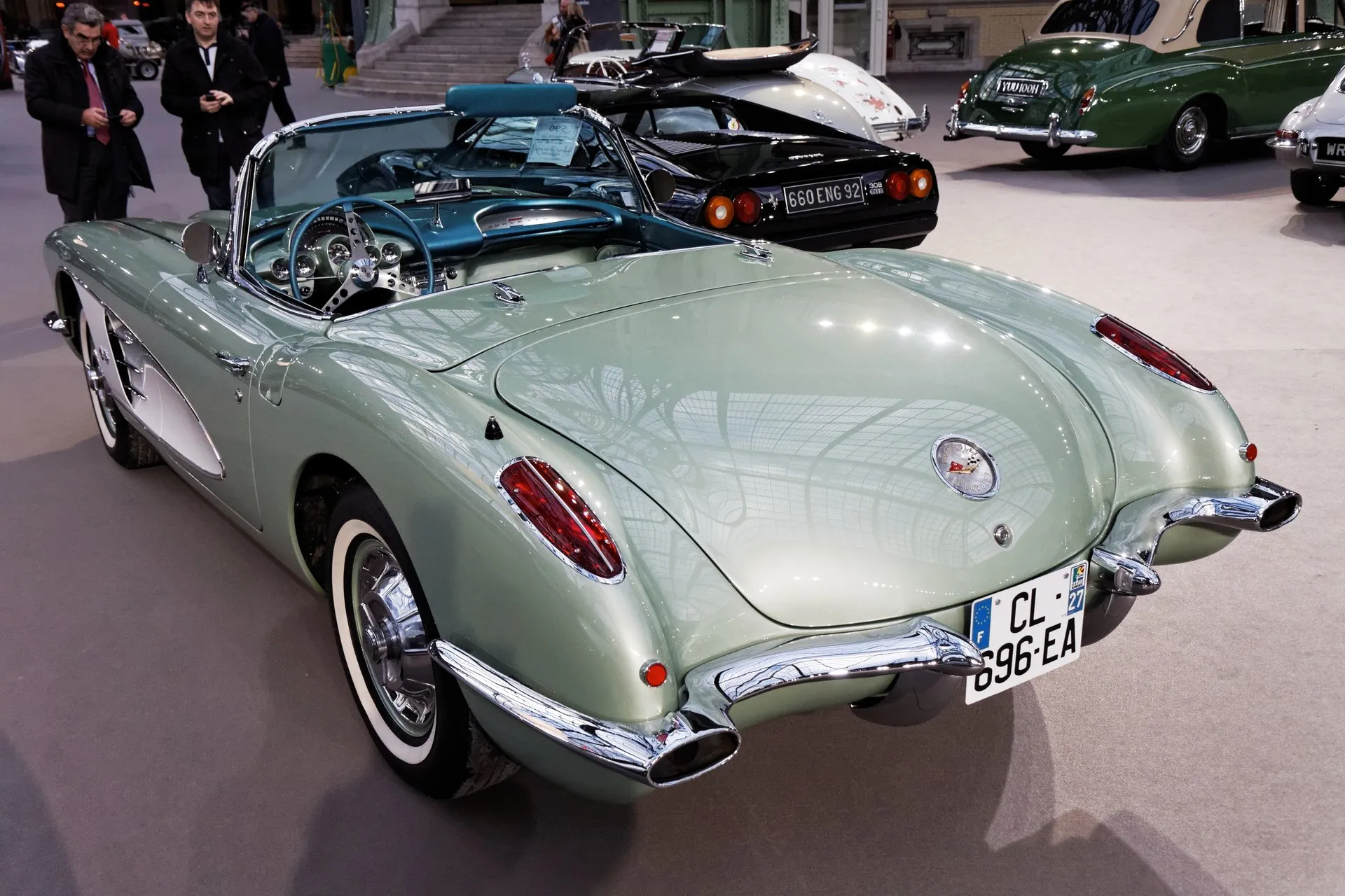 A pale metallic green C1 Corvette with a white side cove and a turquoise interior, seen from behind on an indoor auction floor, showing its taillights set into rounded rear fenders and exhaust tips at the ends of the rear bumper.