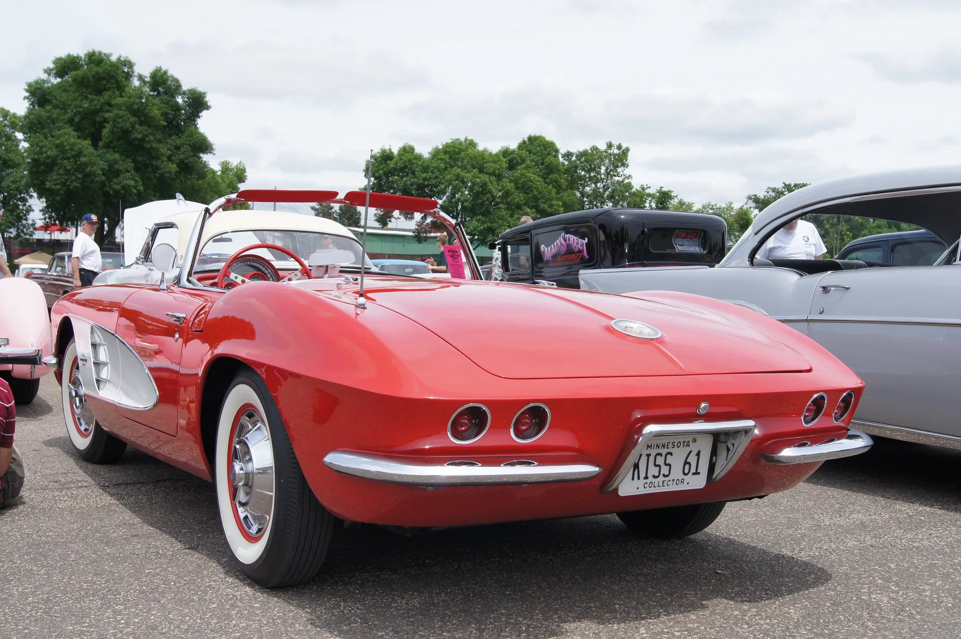 A red C1 Corvette convertible with a white side cove and its top down, seen from the rear left at a car show, showing the flat ducktail rear with four round taillights, a split rear bumper and wide whitewall tires.