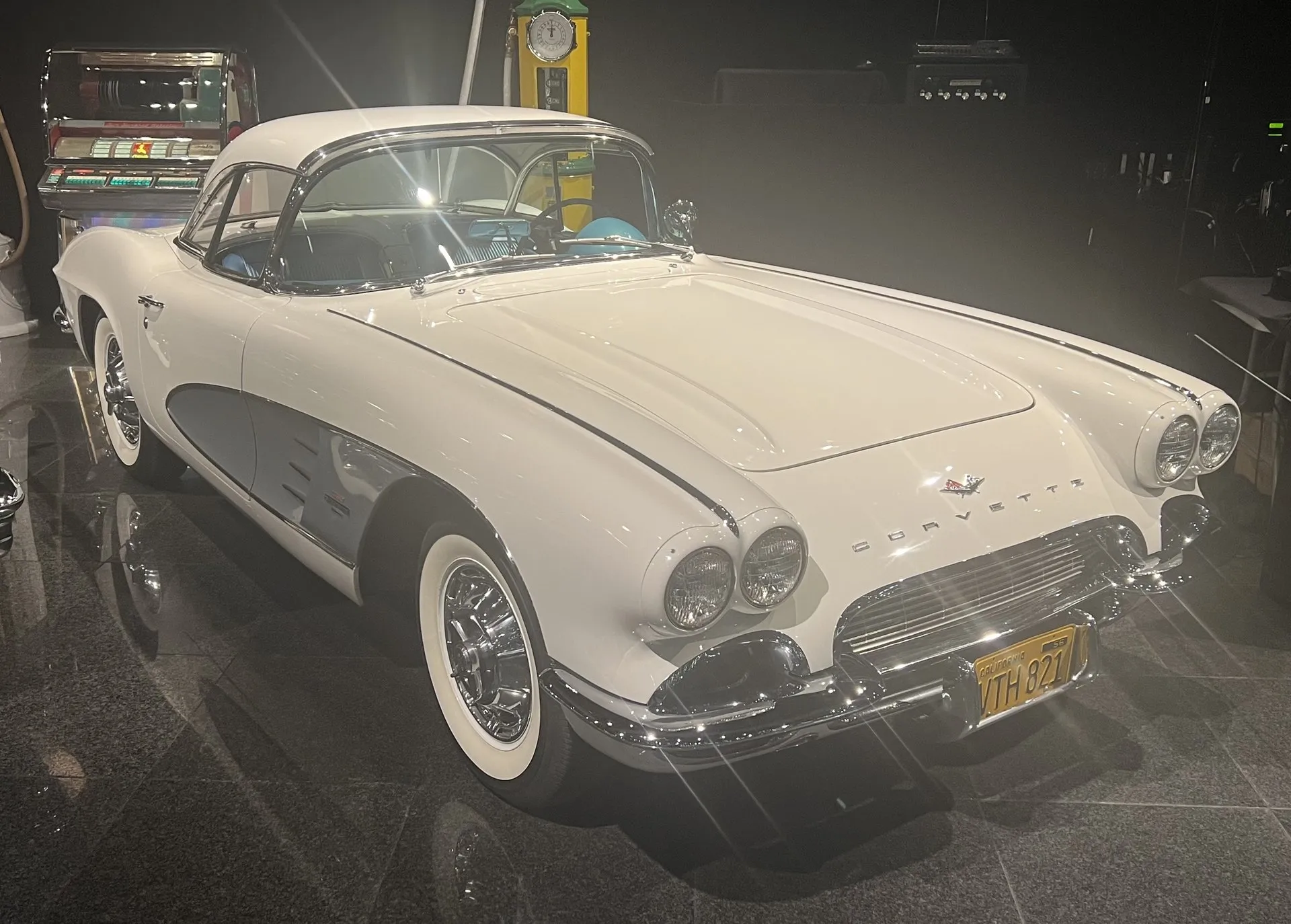 A white C1 Corvette with a silver side cove and a white removable hardtop, displayed on a dark polished floor in a museum beside a jukebox and a vintage gas pump, seen from the front right.