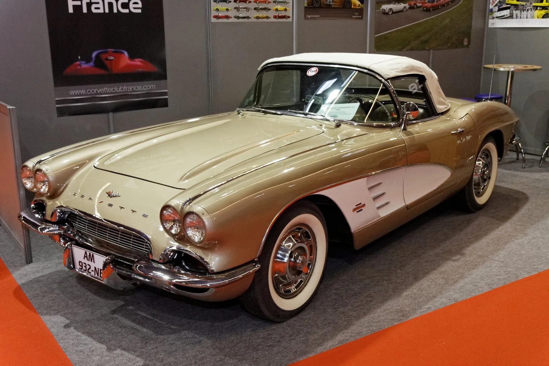 A beige C1 Corvette convertible with a white side cove and a white soft top, on a club stand at an indoor show, seen from the front left, with three bright spears in the cove and wide whitewall tires.