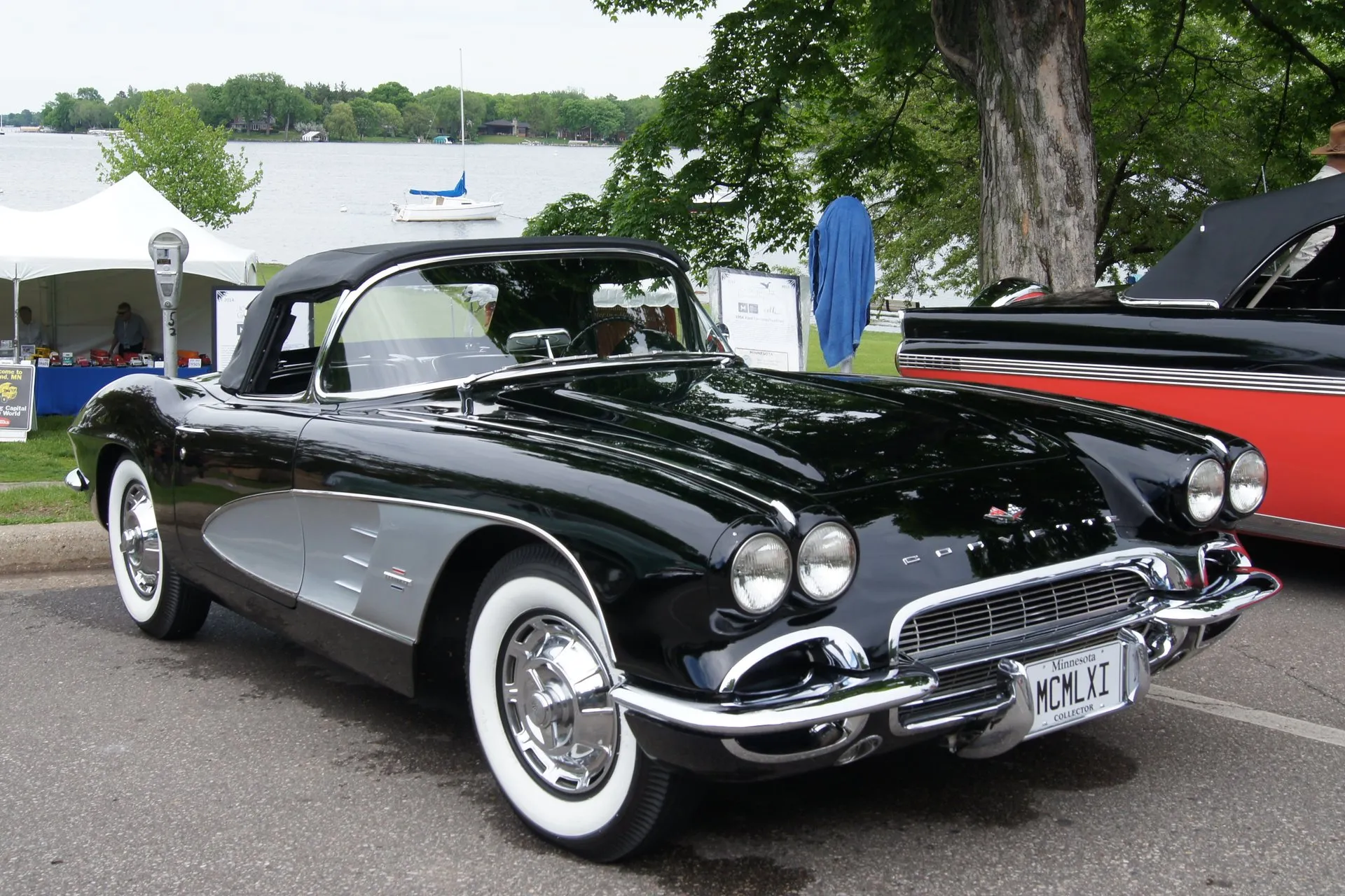 A black C1 Corvette convertible with a silver side cove and its black soft top up, parked by a lake at a car show, seen from the front right, with quad headlights in body-color bezels, a fine mesh grille, wide whitewall tires and full wheel covers.