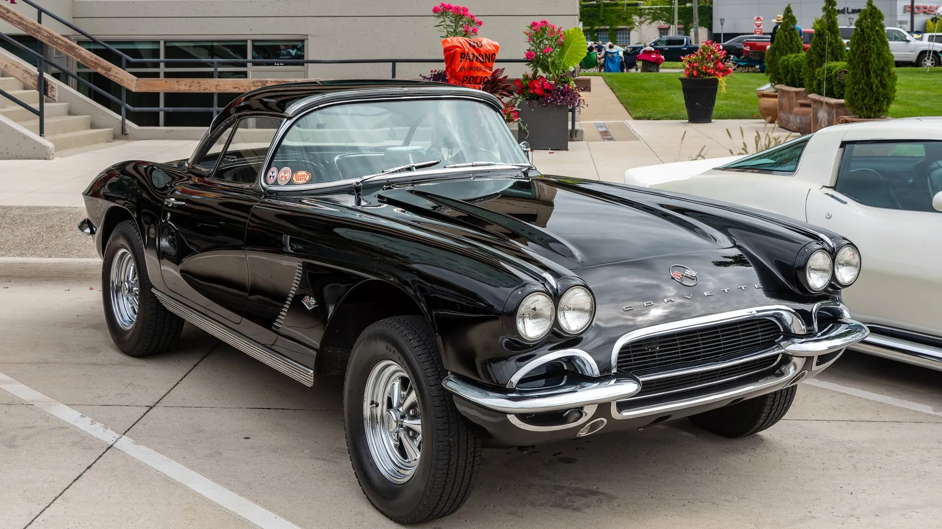 A black 1962 Corvette with a black removable hardtop fitted, parked on a concrete lot and seen from the front right, with a one-color cove, a slatted cove louver, ribbed sill trim and chrome slotted wheels on wide tires.