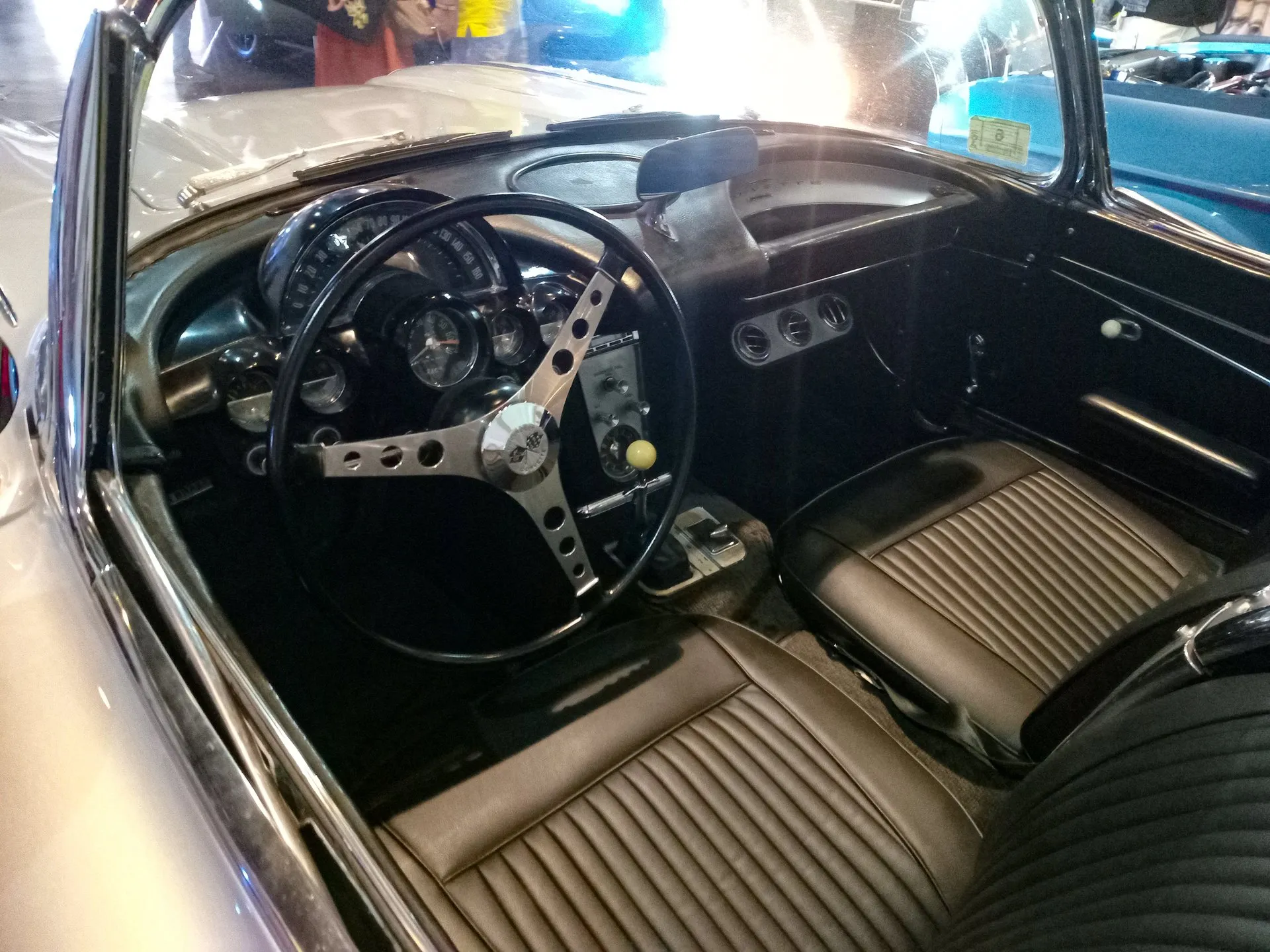 The cockpit of a C1 Corvette convertible with the top down: a speedometer reading to 160 mph arcing over the steering column, round gauges below it, a three-spoke steering wheel with drilled spokes, a floor shifter with a cream knob and brown pleated seats.