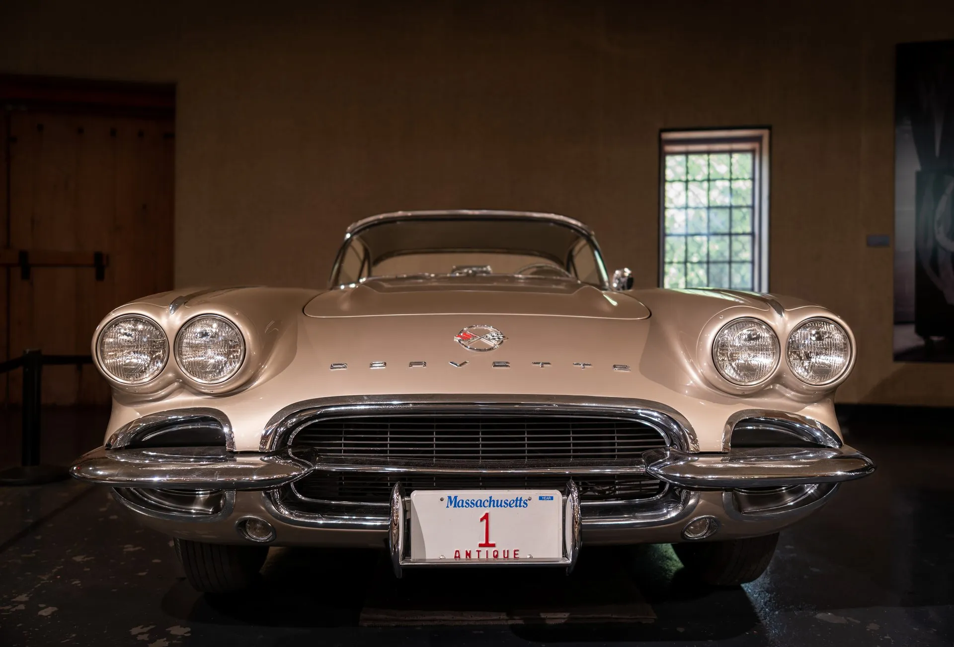 The front of a pale metallic 1962 Corvette in a museum gallery, seen head-on, with four exposed headlights, a crossed-flags emblem above Corvette lettering, a grille of horizontal bars, a split chrome bumper and a Massachusetts antique plate.