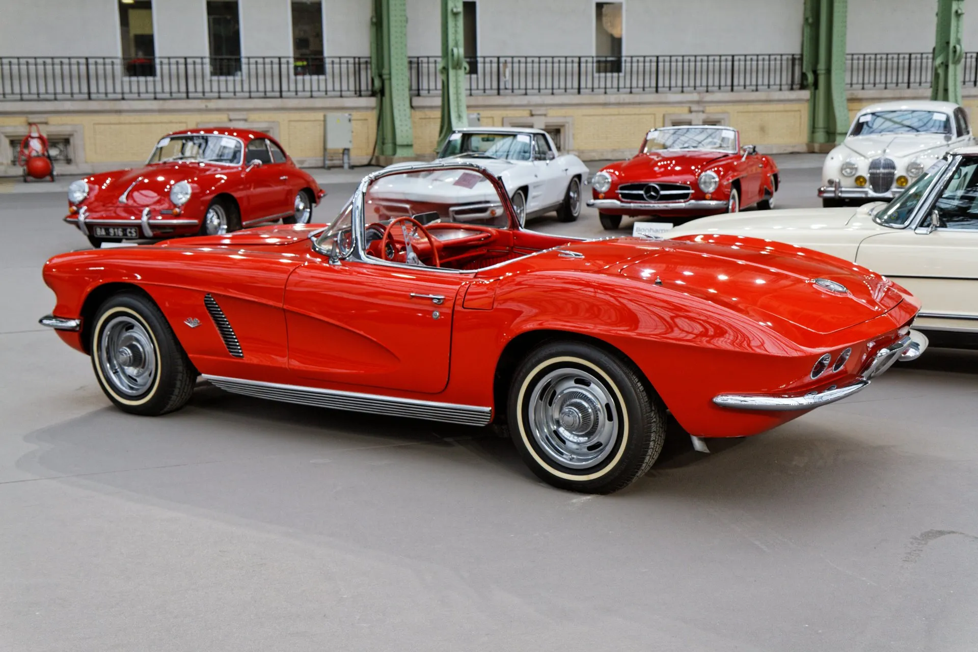 A red 1962 Corvette convertible with its top down on an indoor auction floor, seen from the rear left, showing its trunk lid with a round emblem, the ducktail rear with round taillights, a slatted cove louver, ribbed sill trim and narrow whitewall tires.