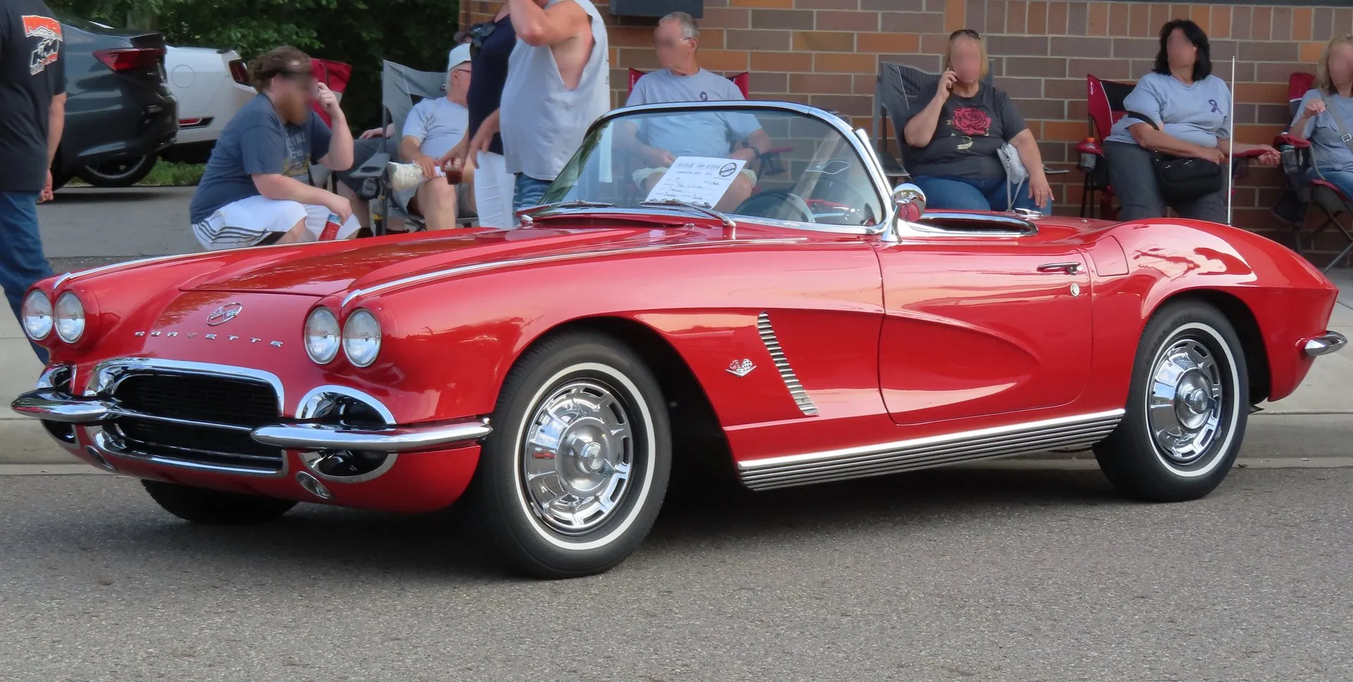 A red 1962 Corvette convertible with its top down and whitewall tires, seen from the front left at a street car show, with one-color side coves, a slatted louver in the cove and ribbed bright trim along the sill. Spectators sit in folding chairs behind it, their faces blurred.