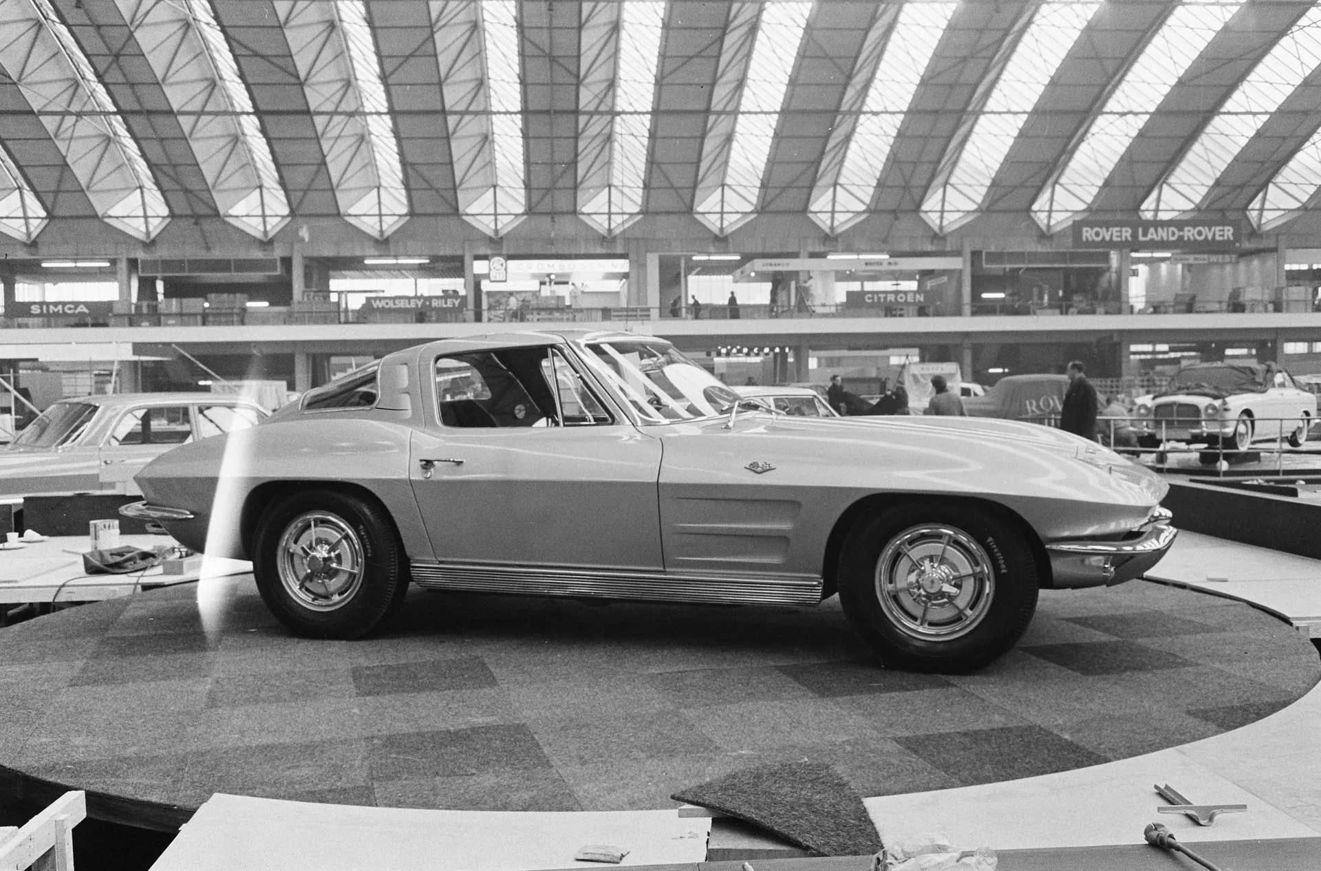 A black-and-white photograph of a Corvette Sting Ray coupe in side profile on a round display stand at an indoor motor show, under an arched glass roof, with Rover Land-Rover, Citroën and Simca signs on the balcony behind it and workers setting up stands.
