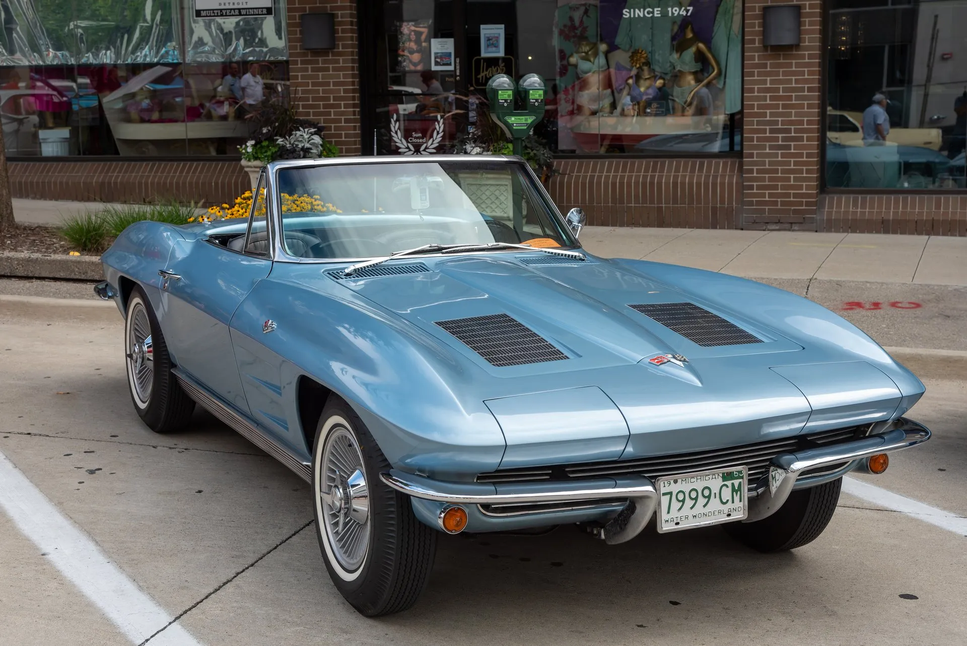 A light blue 1963 Corvette convertible with its top down, parked at a curb in front of a shop and seen from the front right, with two trim panels in the hood, whitewall tires and a 1963 Michigan license plate.