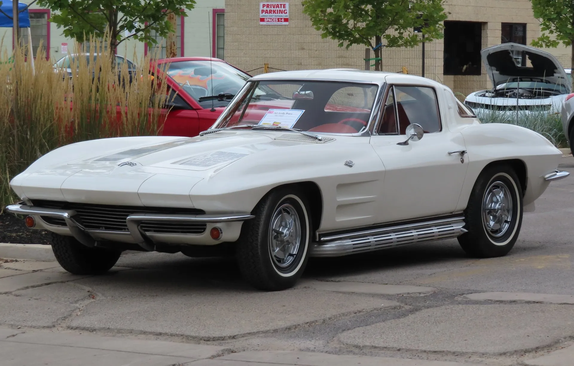 A white 1963 Corvette Sting Ray coupe with a red interior, parked in a lot at a car show and seen from the front left, with two rectangular trim panels set into the hood, ribbed chrome rocker trim and whitewall tires on full wheel covers.