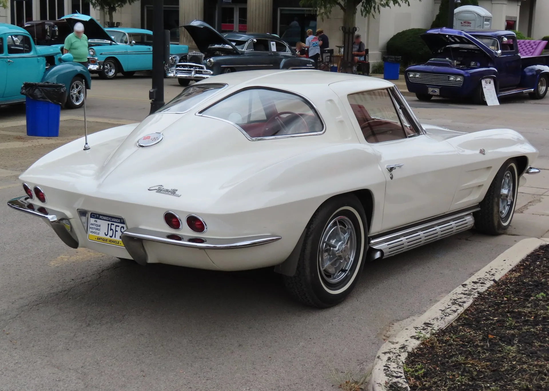 The same white 1963 Corvette coupe seen from the rear right at a car show, with a body-color spine dividing the rear window into two panes, round tail lights and a Pennsylvania antique vehicle plate.