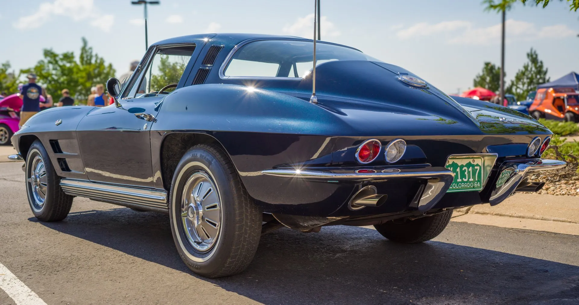 A dark blue 1964 Corvette coupe at an outdoor car show, seen from the rear left, with a one-piece rear window, louvered vents in the roof pillar behind the door, two horizontal vents in the front fender and a green Colorado 64 license plate.