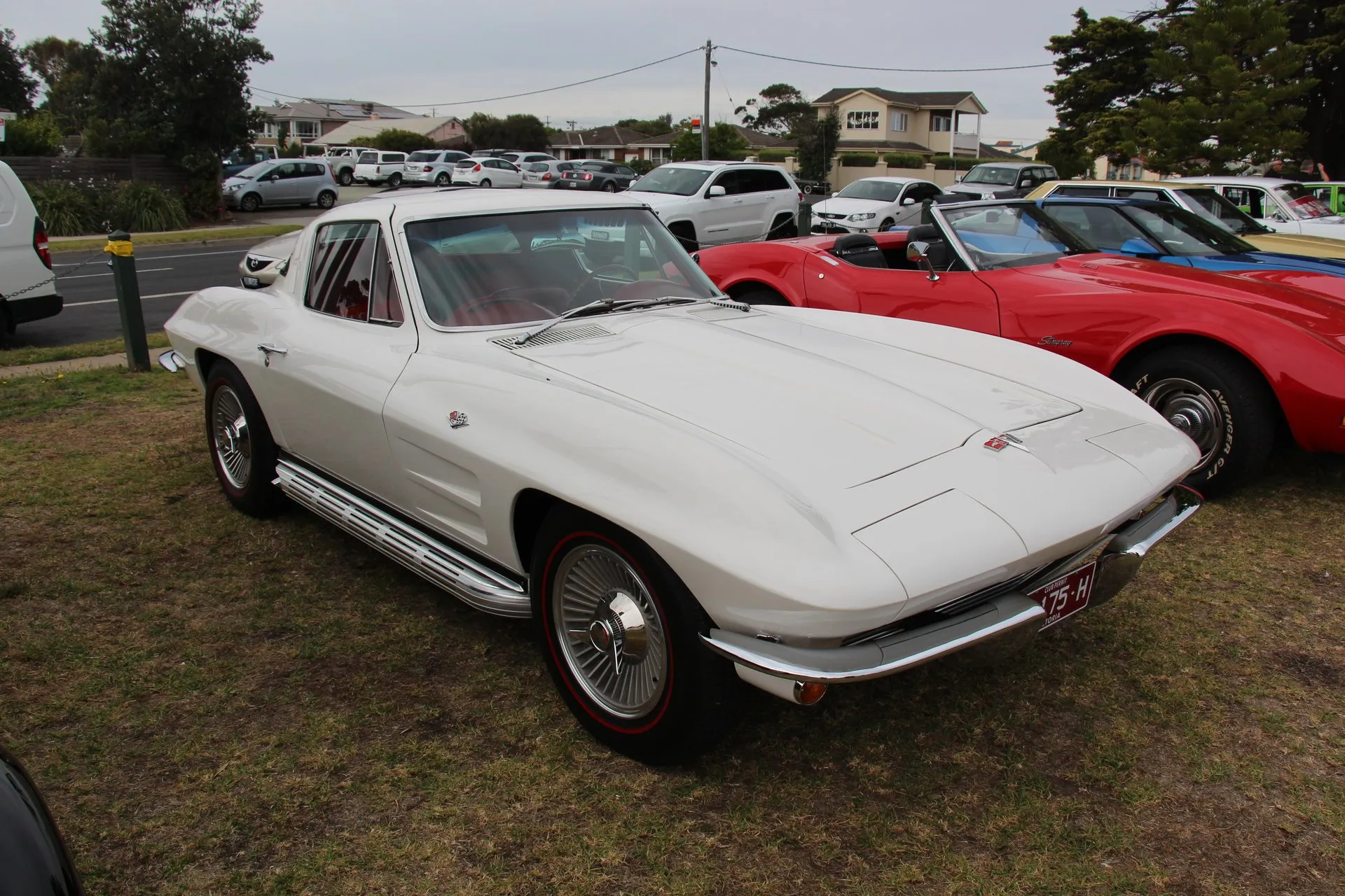 A white 1964 Corvette coupe with a red interior parked on grass at a car show, seen from the front left, with two long recesses in the hood, two horizontal fender vents, ribbed rocker trim and wheel covers on red-stripe tires.